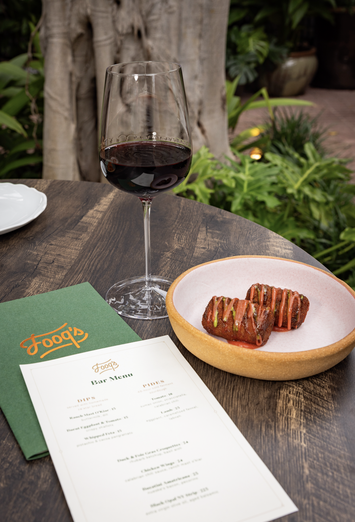 A glass of red wine and two pieces of fried food with sauce on a pink plate on a wooden table outside. There is also a green menu, a white paper, a white plate, and green plants in the background.