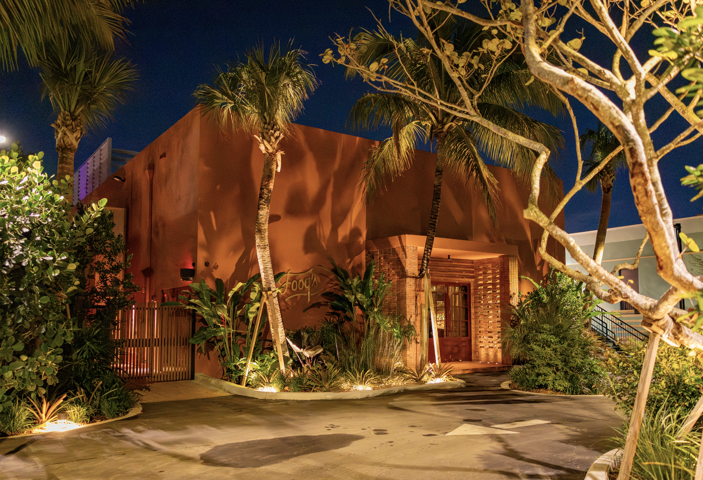 Nighttime scene of a tropical courtyard with palm trees and various plants illuminated by warm lighting, with a building in the background that has a sign reading 'Food's'.