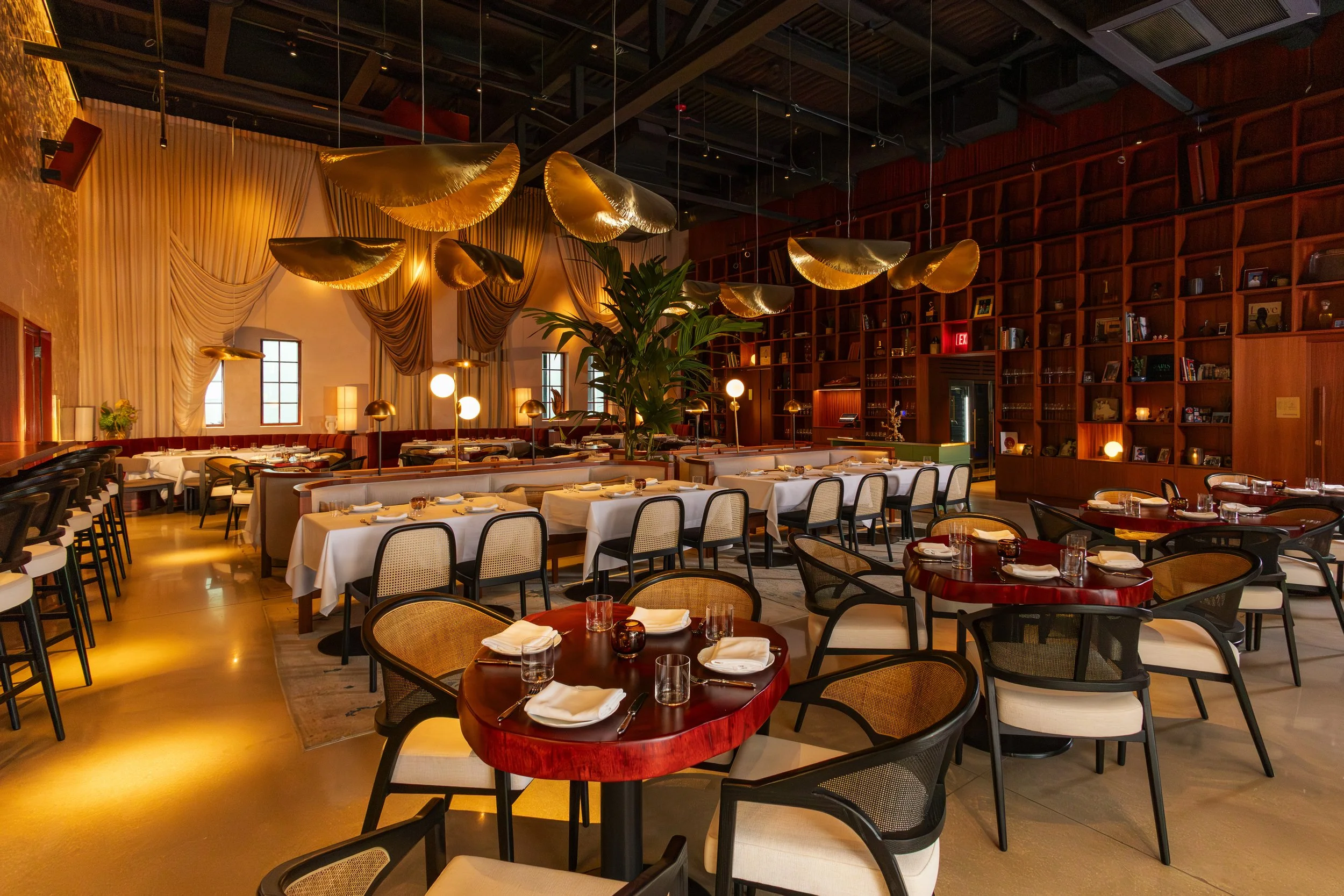 Elegant restaurant interior with round wooden tables set with napkins, glasses, and small candles, surrounded by black and beige chairs. Golden leaf-shaped light fixtures hang from a dark ceiling. A large wooden bookshelf wall, draped curtains, and t