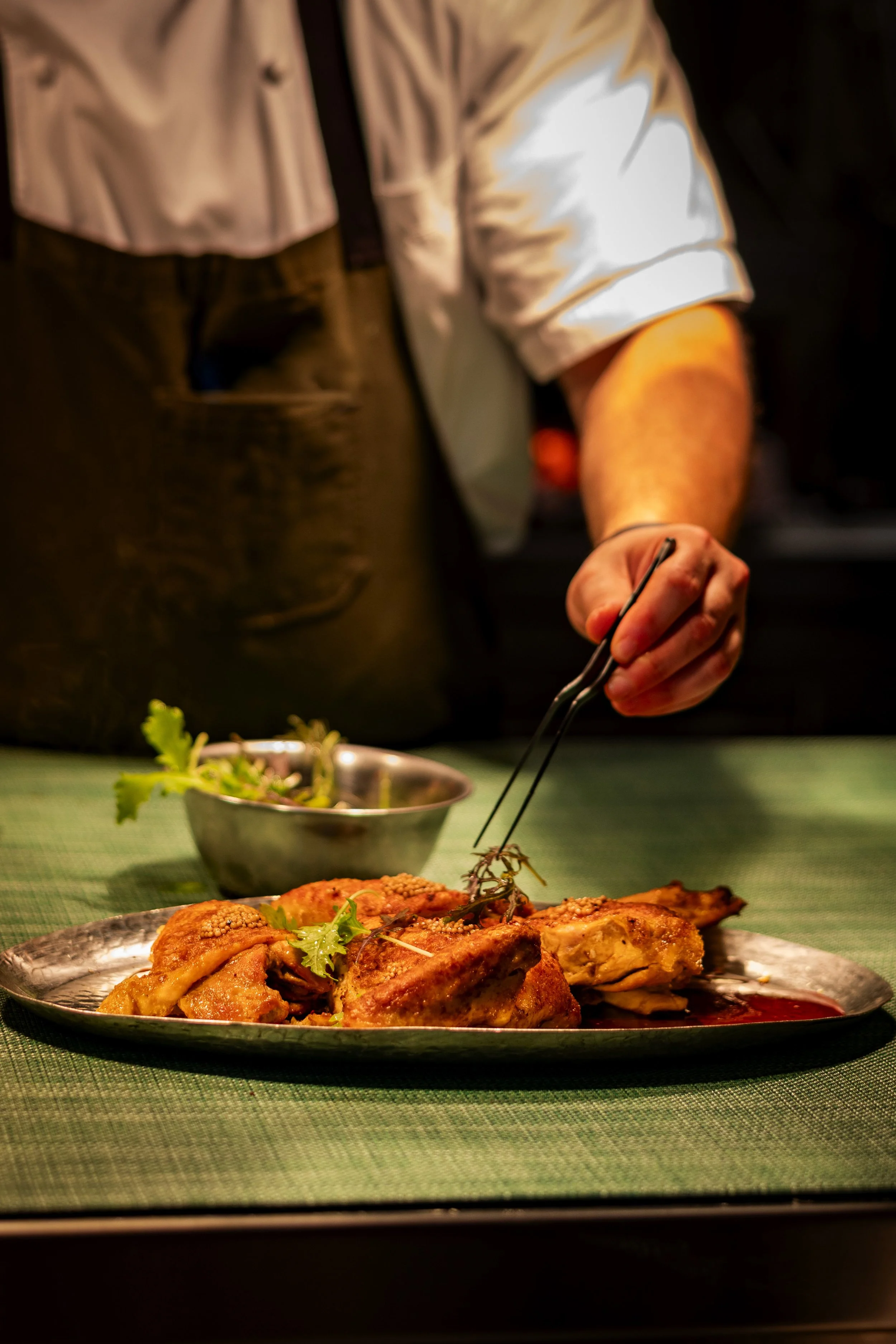 A person serving food onto a silver plate of cooked chicken garnished with herbs, with a bowl of salad in the background.