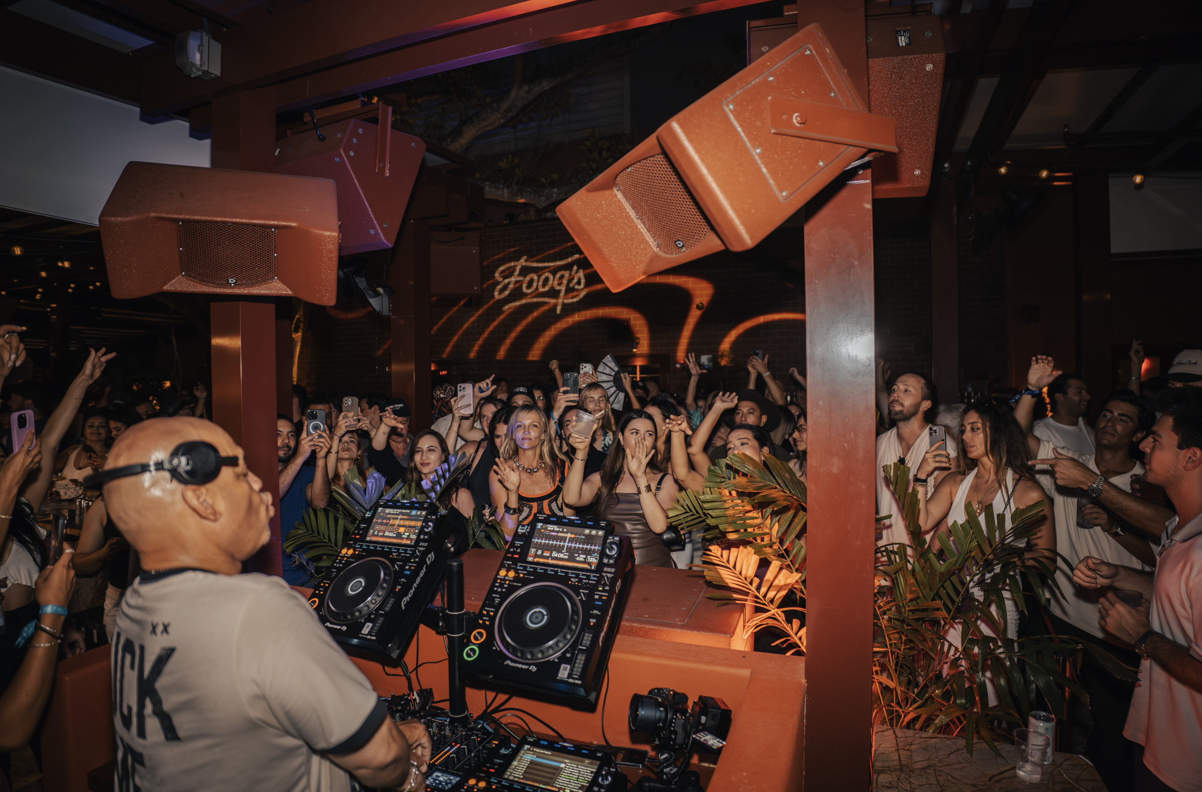A DJ wearing a beige t-shirt with black text is performing at a crowded nightclub with a tropical decor theme. The scene is lively with many people dancing and holding up drinks, illuminated by orange lighting.