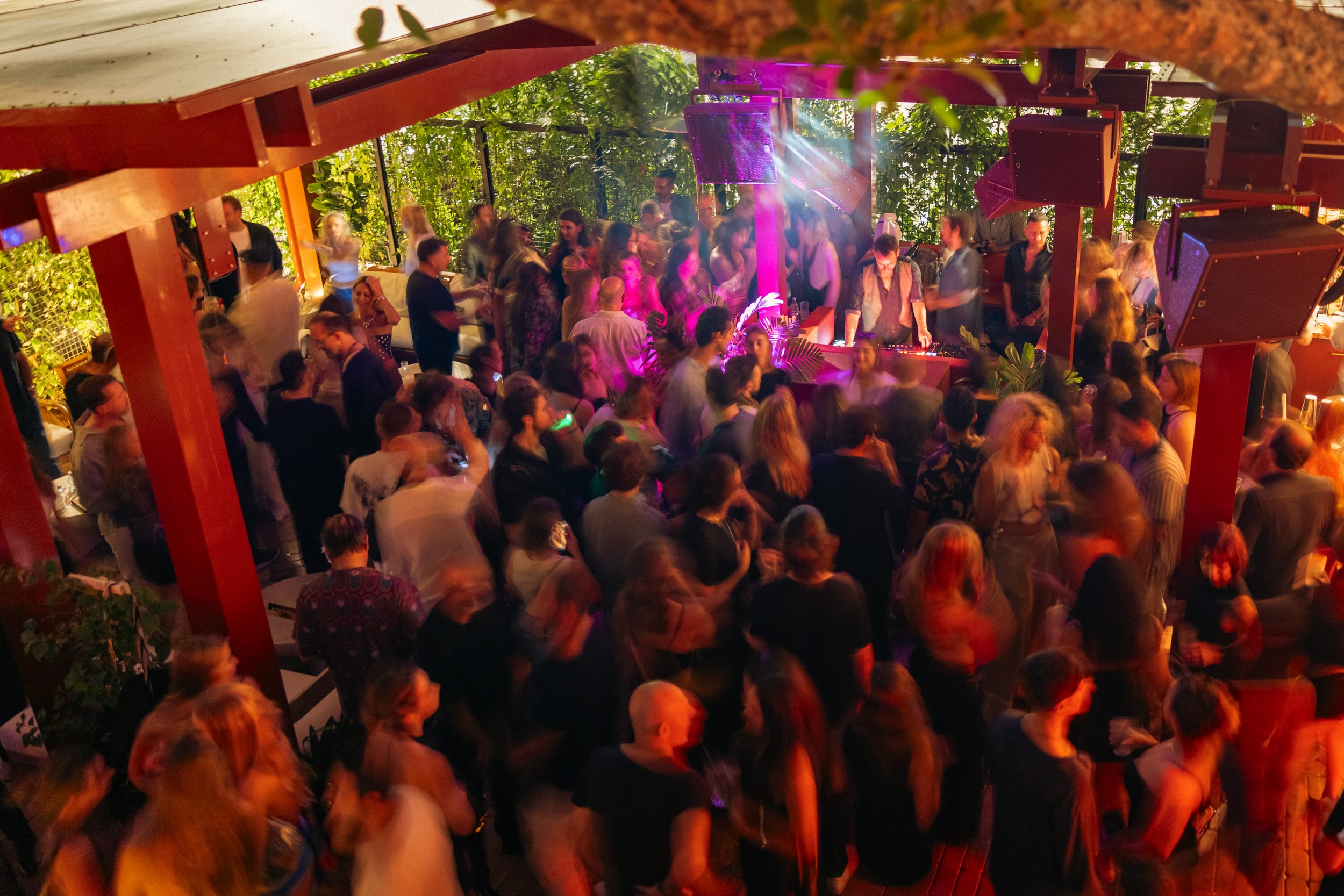 A crowded dance floor with people dancing, a DJ playing music, and stage lighting in a vibrant, lively venue with green leafy decor.