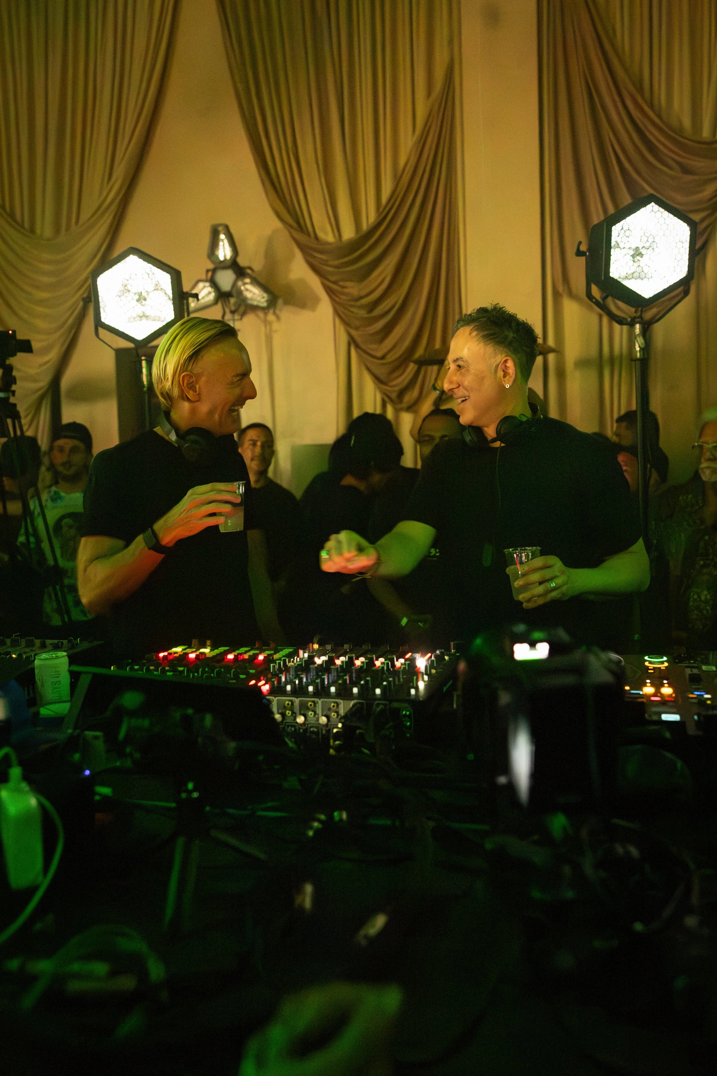 Two people, a man with blonde hair and a woman with short hair, are smiling and talking at a party or event, with DJ equipment and lighting around them.