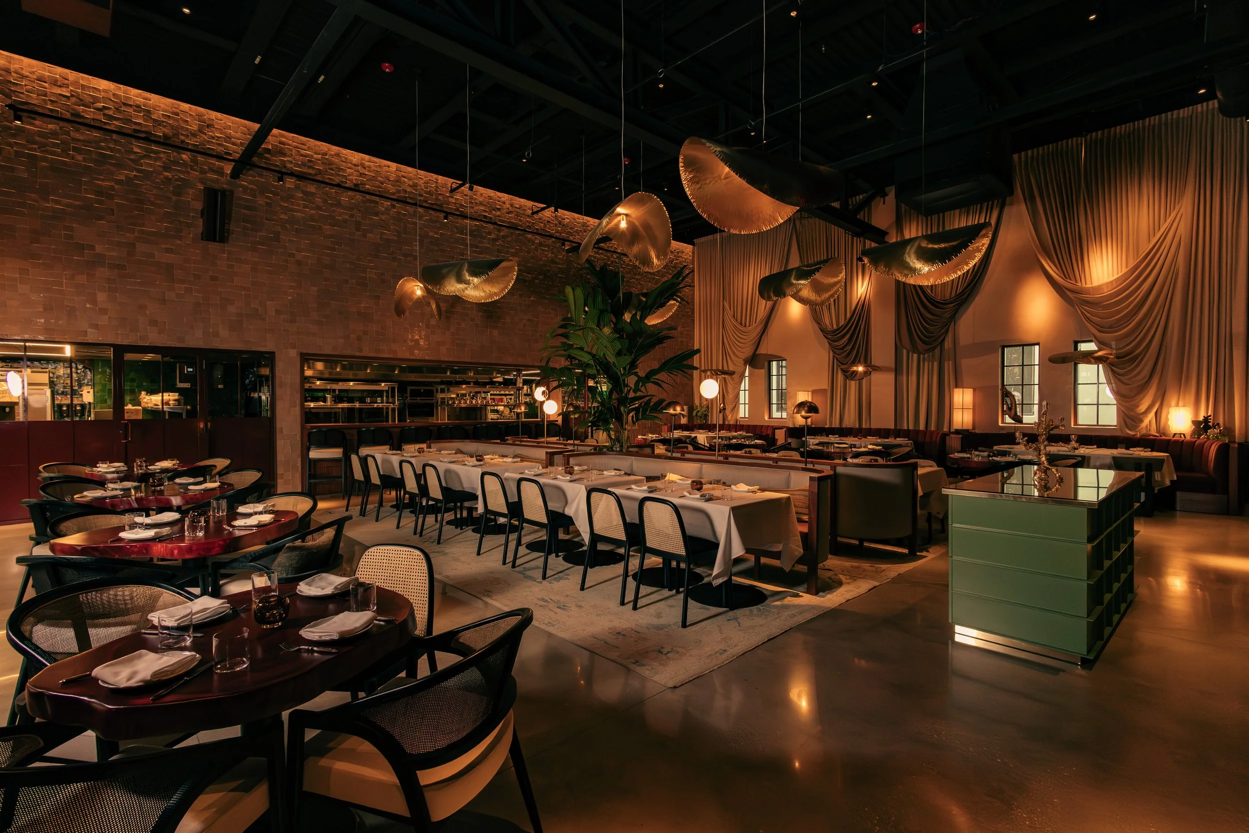 Elegant restaurant interior with tables set with white tablecloths, glassware, and napkins, warm lighting, and decorative gold hanging light fixtures, brick and draped curtains decor, and large windows.