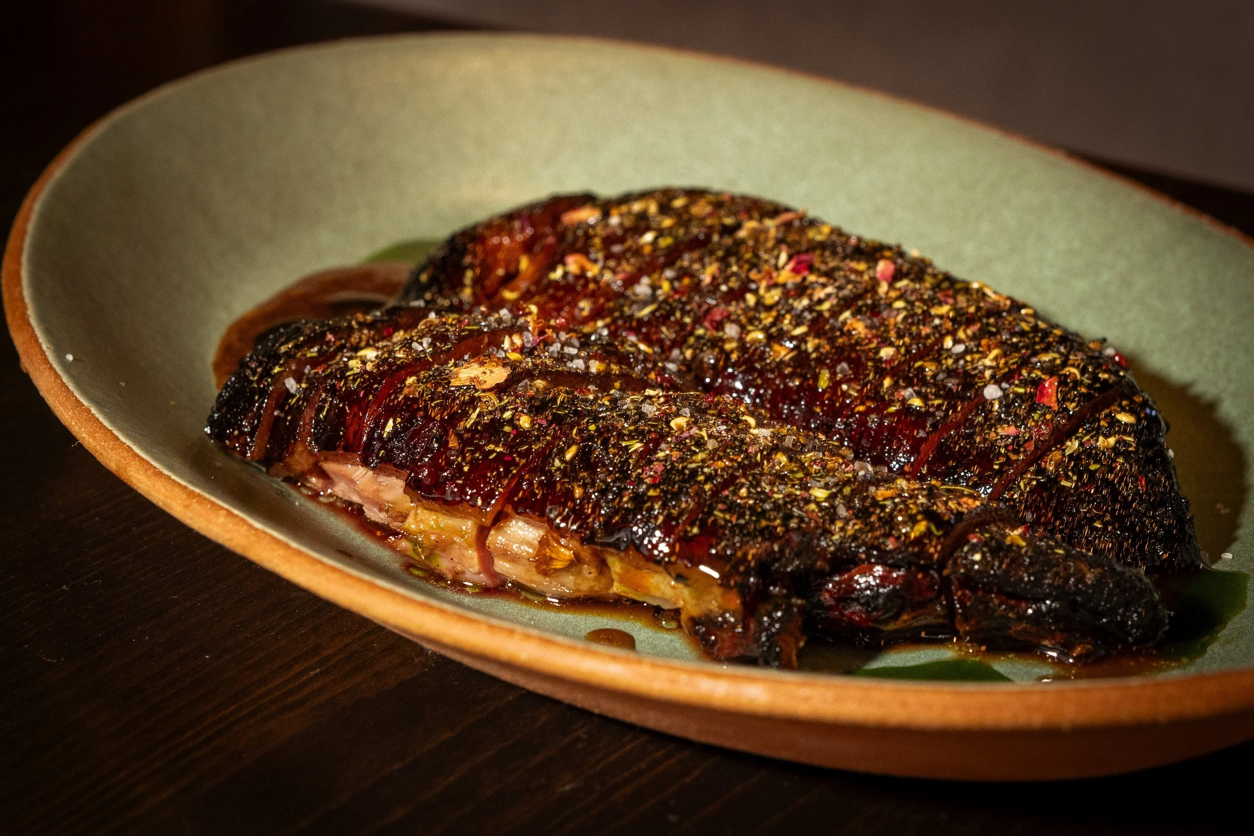 A cooked meat roast with a black pepper crust, resting on a green leaf in a ceramic dish.
