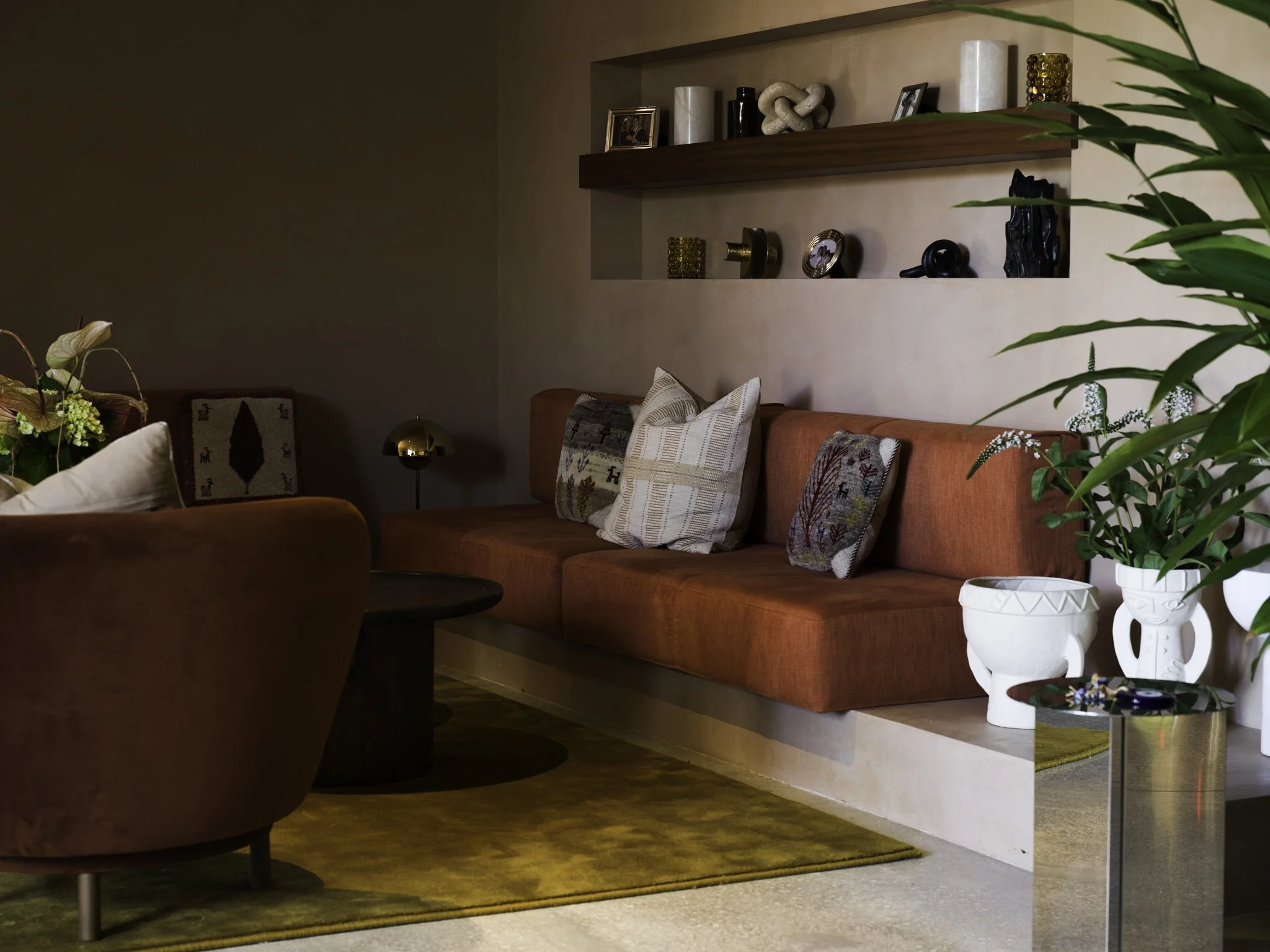 Living room with a peach-colored sofa, patterned pillows, a white decorative planter with a large green plant, a small round black table, a yellow rug, a wall shelf with decorative vases, photo frames, a black sculpture, and a gold accent table benea