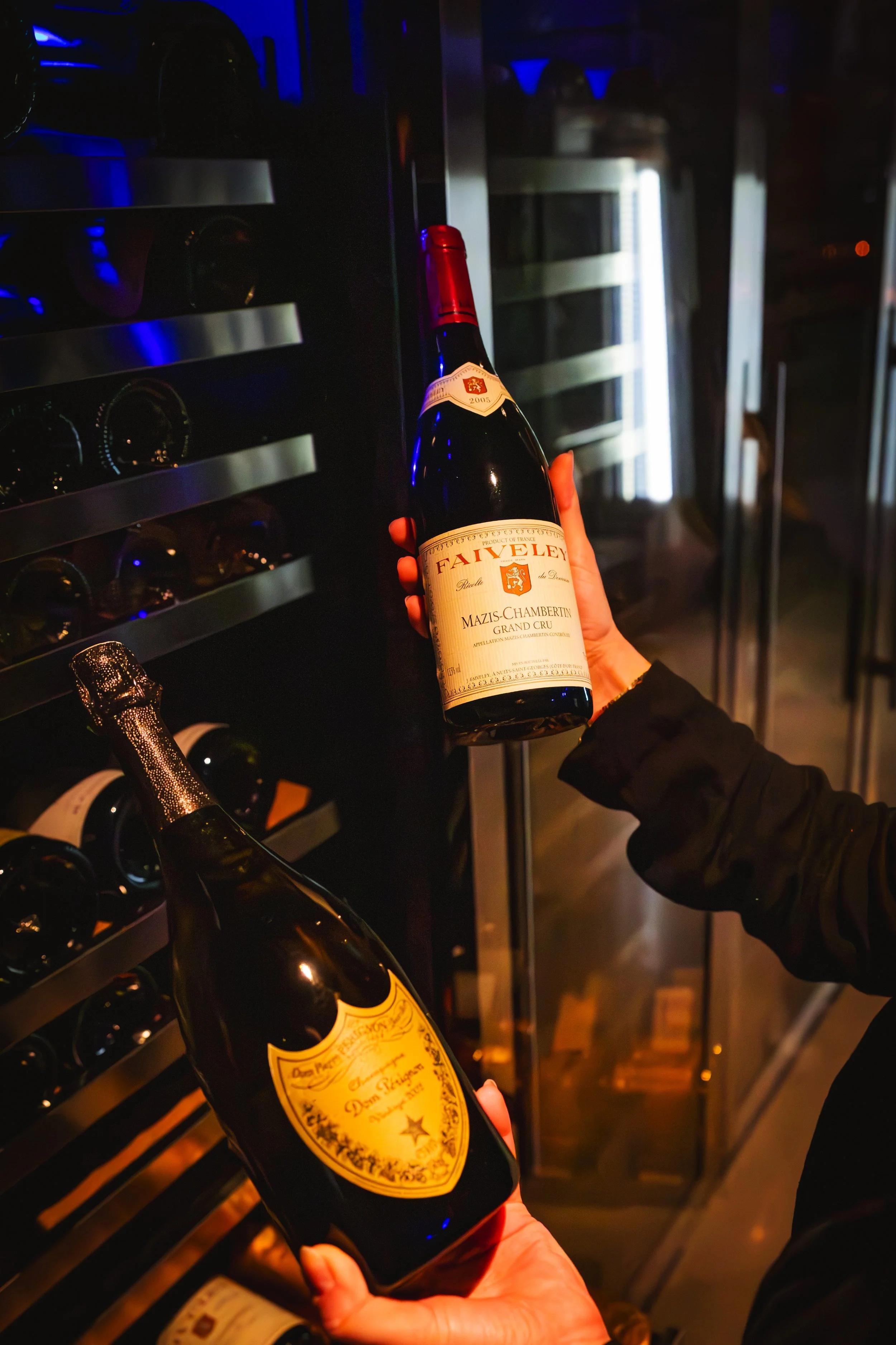 Person holding a bottle of red wine in front of a wine cooler filled with bottles in a dimly lit setting.