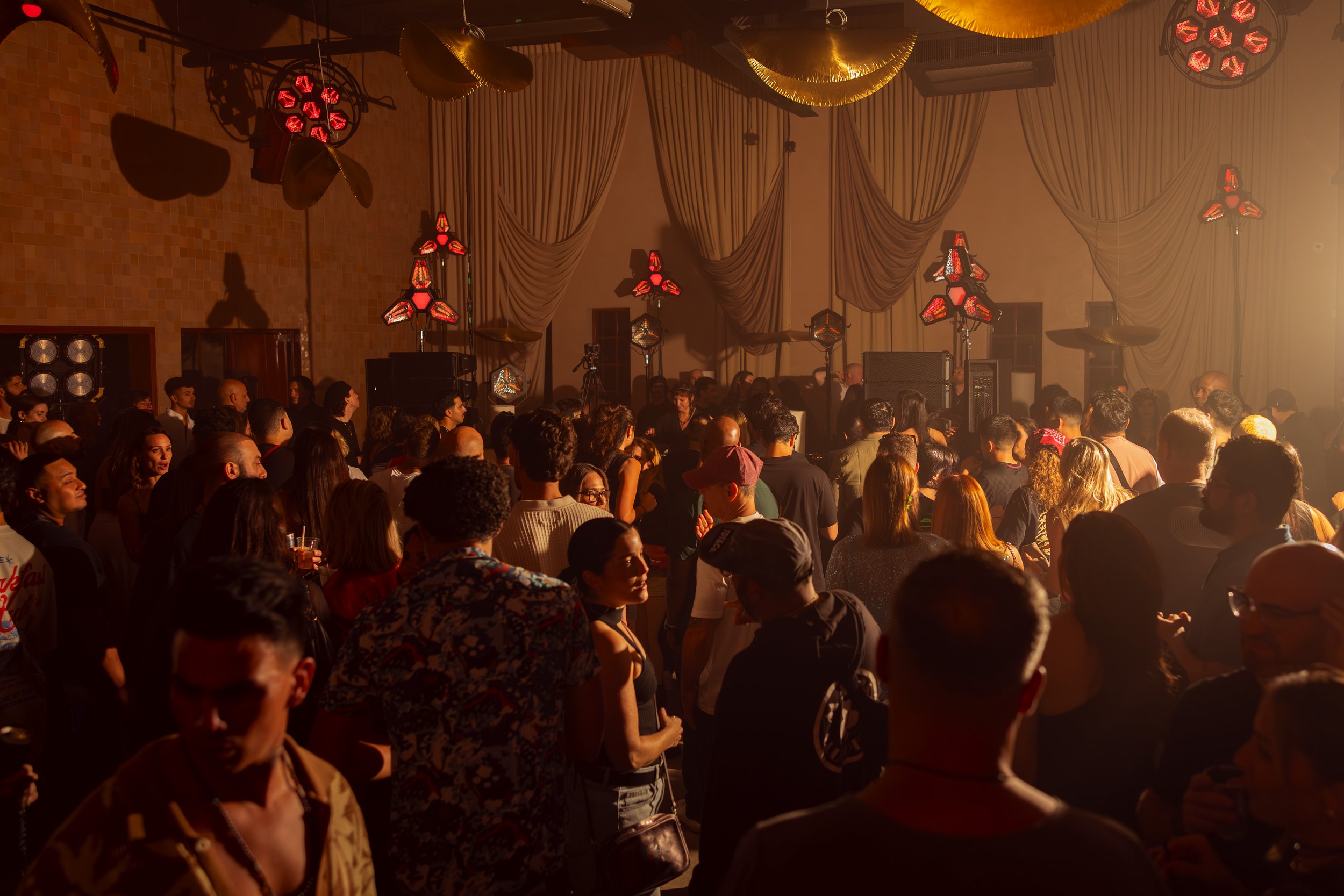 Crowd of people at an indoor concert or event with stage lighting and decorative hanging lights.