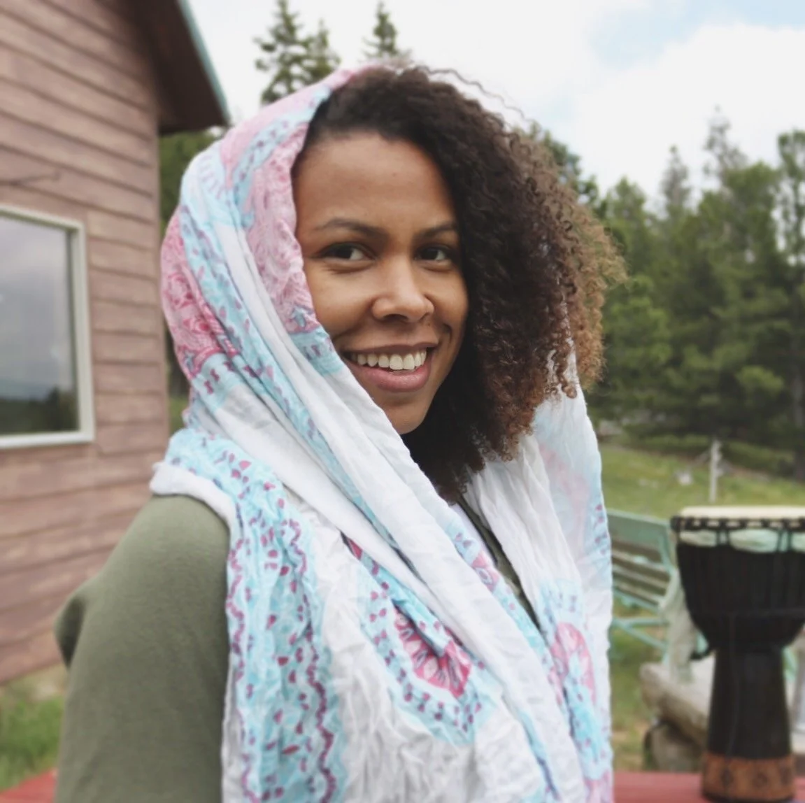 Smiling woman outdoors with a colorful headscarf, green hoodie, wooden house, trees, and a drum in the background.