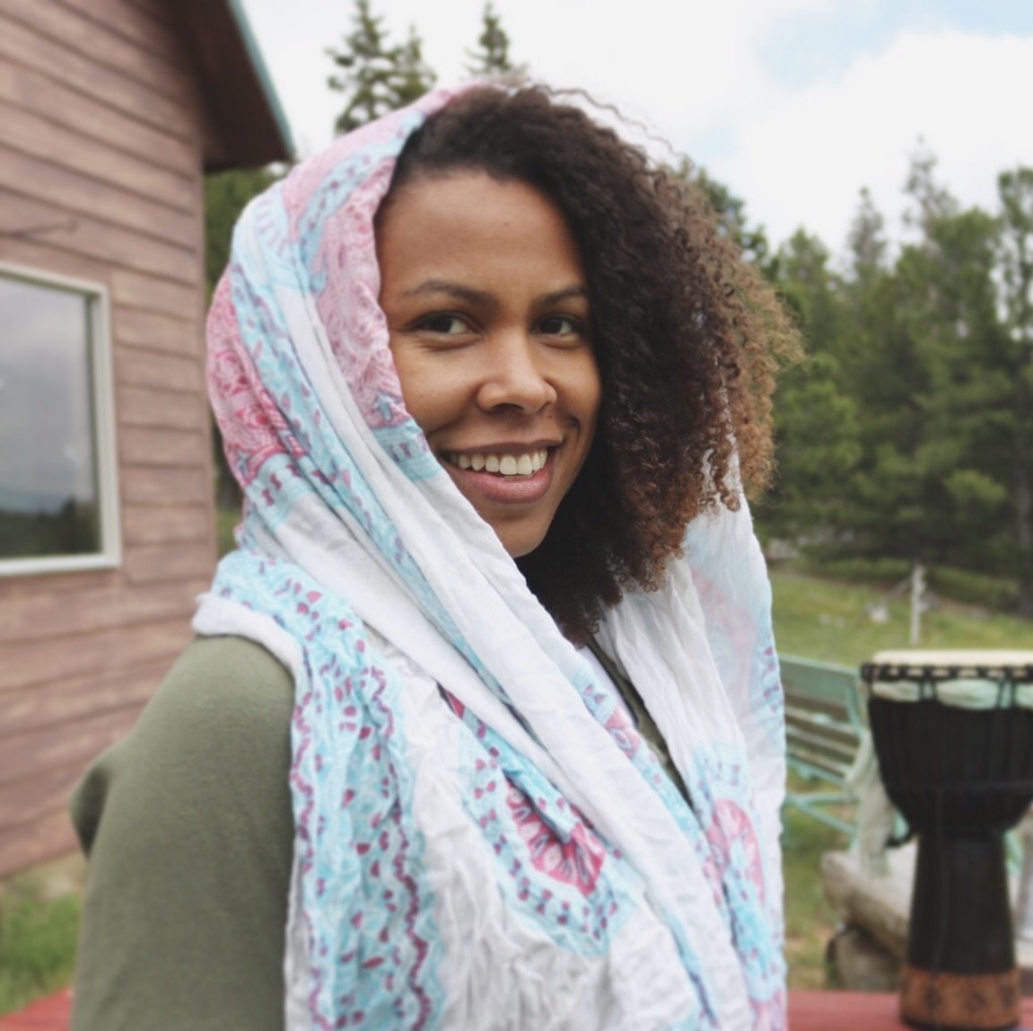 A woman smiling outdoors with a colorful scarf over her head, standing near a wooden building and greenery.