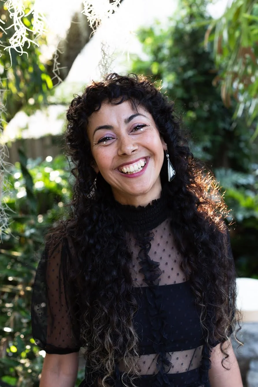 A woman with curly black hair smiling outdoors, wearing a black sheer polka dot top and earrings, surrounded by greenery.