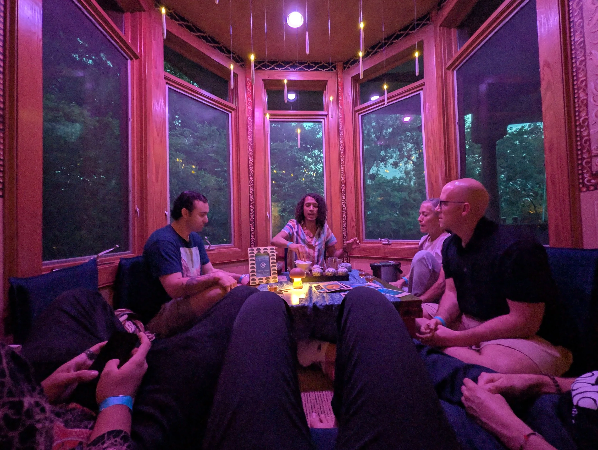 A group of five people sitting around a table in a room with large windows and purple lighting. The people appear to be engaged in conversation, with some holding hands, and a woman in the center speaking. The table has various items, including a tarot card deck, candles, and drinks.