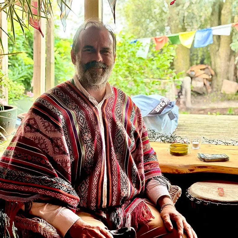 A man with a beard and gray hair sitting at a wooden table on a porch, wearing a red, black, and white patterned poncho, with a smile, surrounded by greenery, colorful flags, and decorative items on the table.