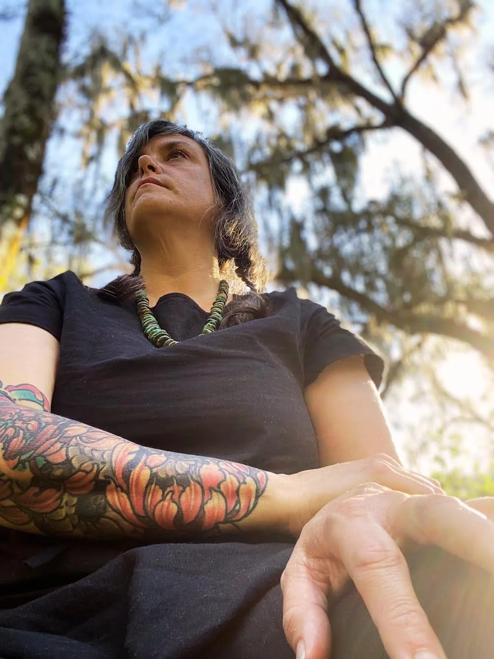 A woman with dark hair and tattoos on her arm sitting outdoors under a tree, looking up at the sky.
