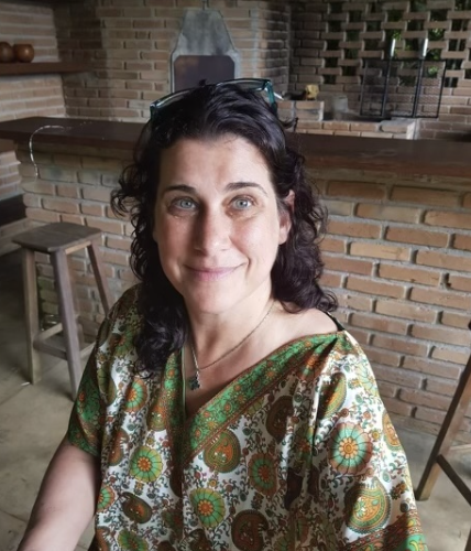 A woman with dark curly hair wearing a colorful patterned blouse, sitting in a cozy brick-walled cafe or restaurant with a wood bar and barstools in the background.