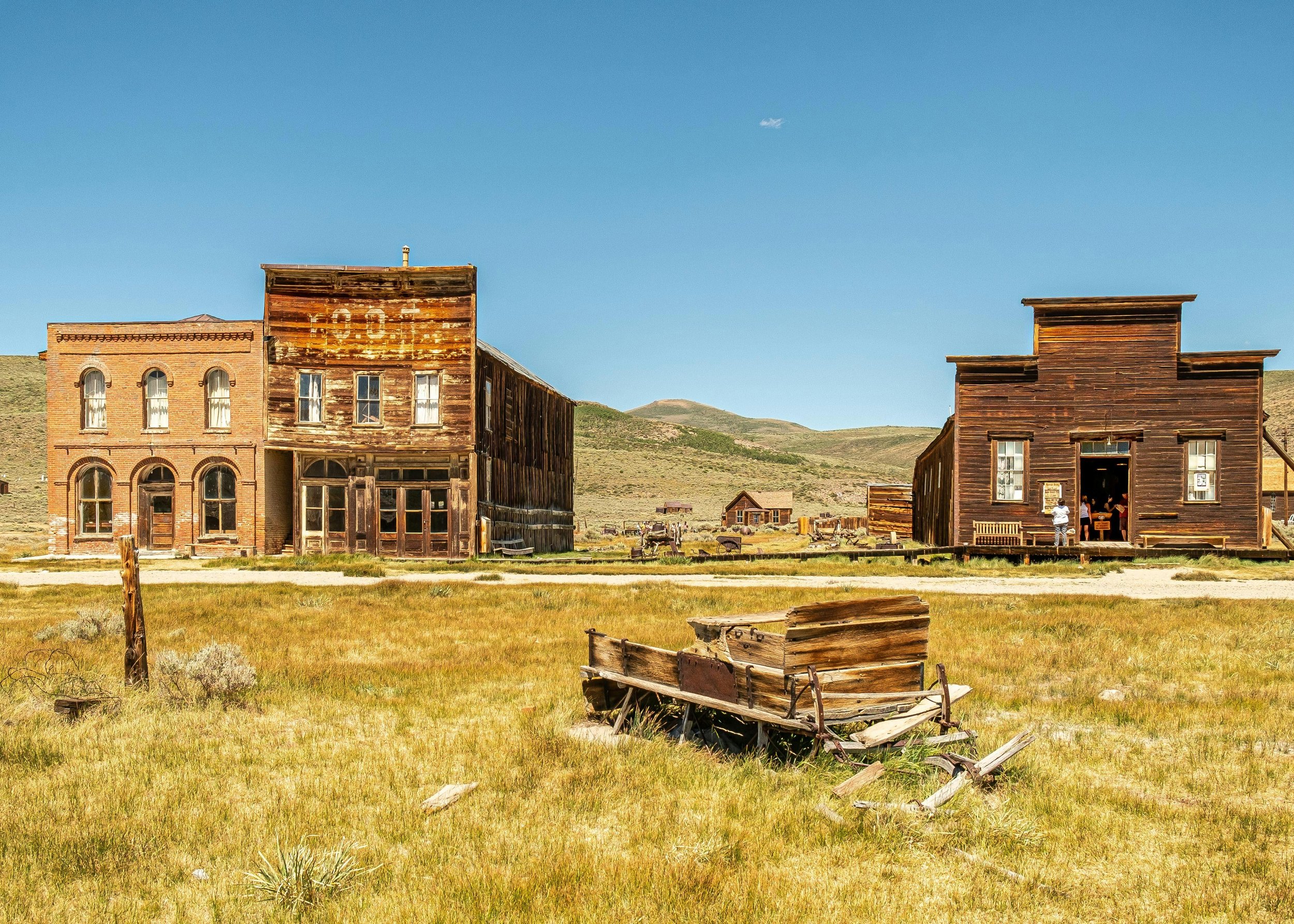 Old Western town scene featuring weathered wooden buildings, some with broken windows, amid a grassy plain under a clear blue sky.