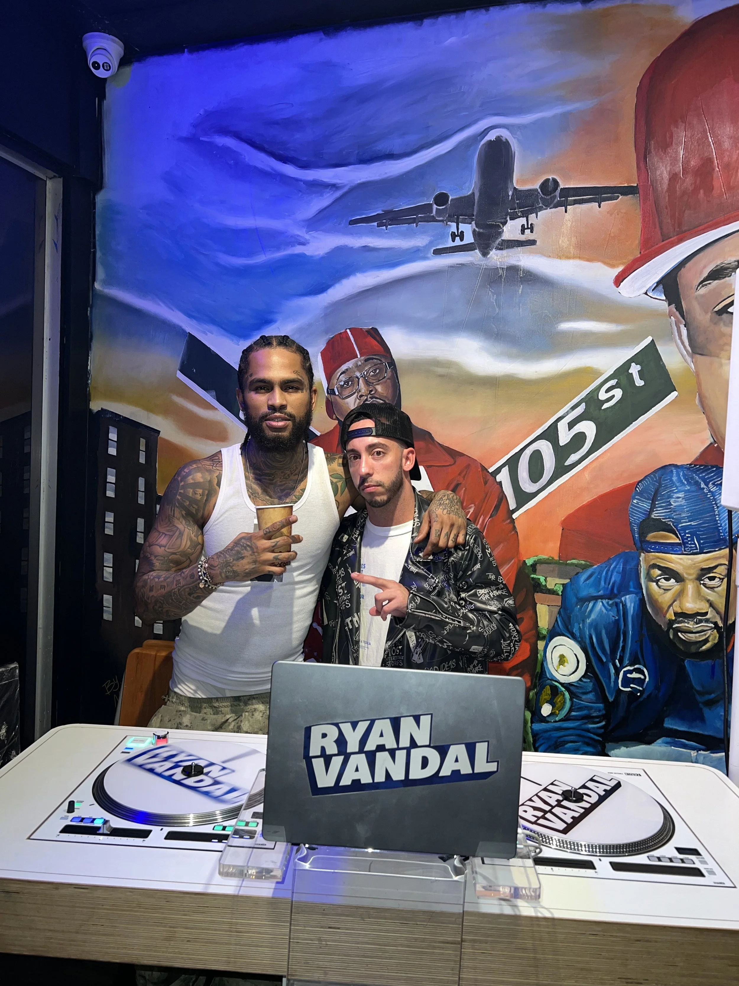Two men pose in front of a colorful mural with an airplane and rapper Tupac Shakur. There is DJ equipment on a table with a sign reading 'Ryan Vandal'.