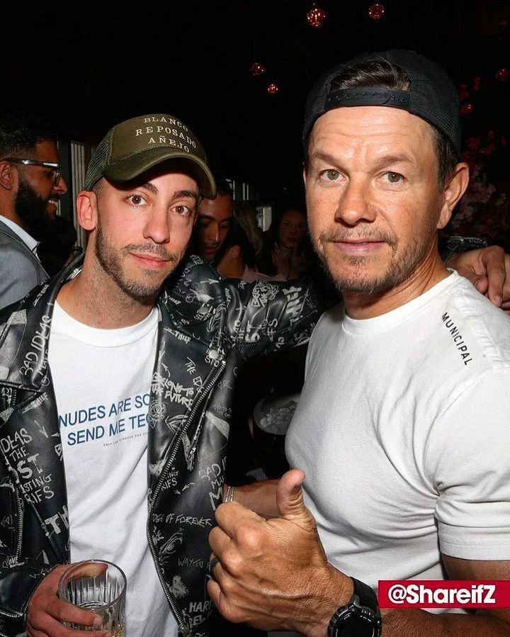 Two men posing for a photo at a social event. The man on the left wears a baseball cap, a graphic jacket, and a white T-shirt, holding a drink. The man on the right wears a backwards cap, a white T-shirt, and gives a thumbs-up. Background shows other people and hanging lights, suggesting an indoor party or celebration.