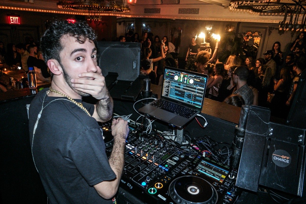 DJ performing at a crowded nightclub with a laptop, mixing equipment, and a lively audience.