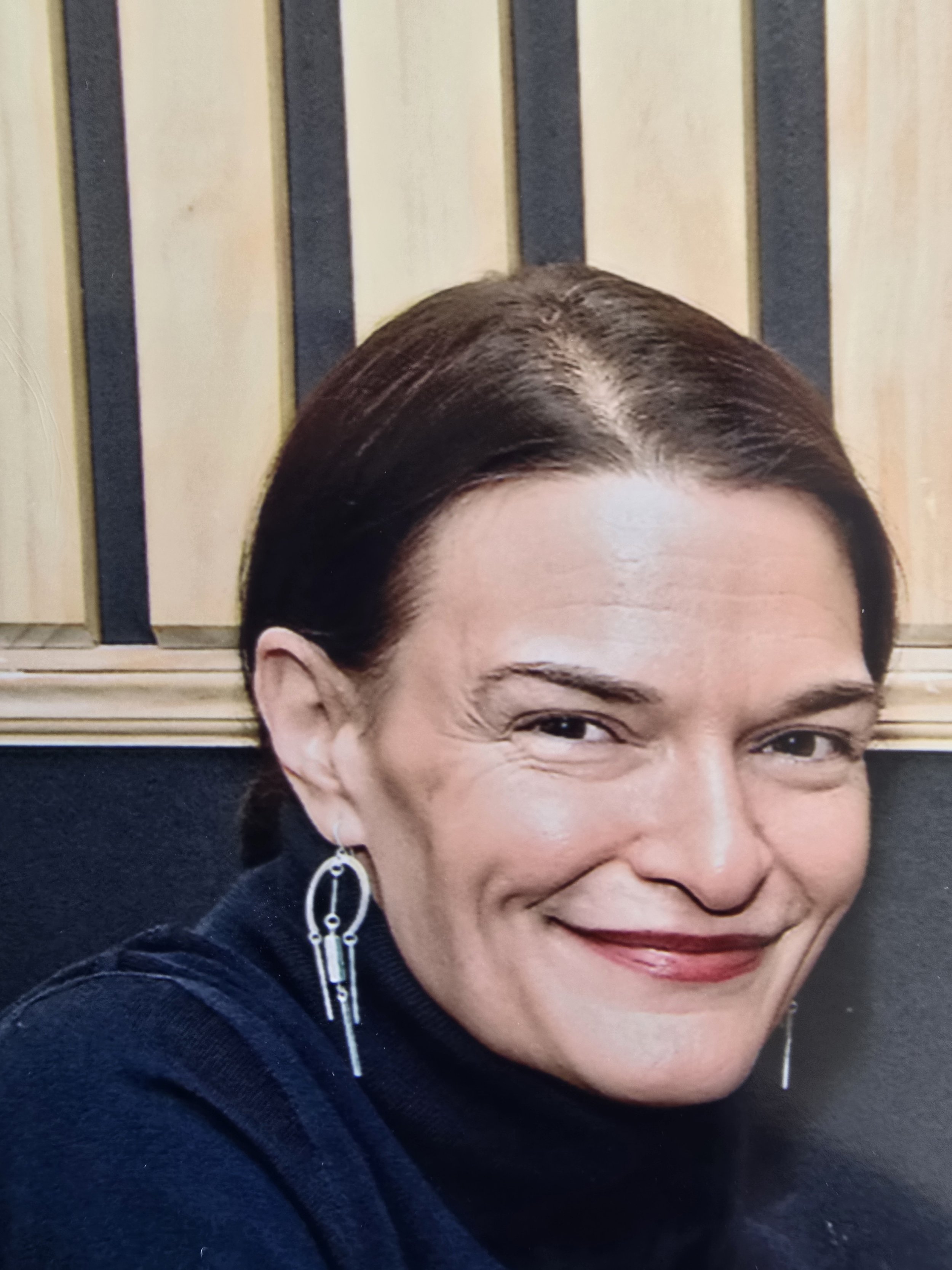 Close-up of a smiling woman with dark brown hair styled in a bun, wearing earrings and a dark top, standing in front of a wooden background with black vertical stripes.