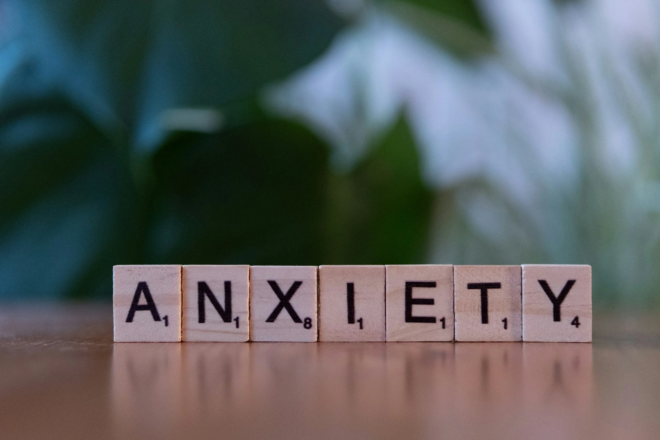 Scrabble tiles spelling out 'ANXIETY' on a wooden surface with a blurred green leafy background.