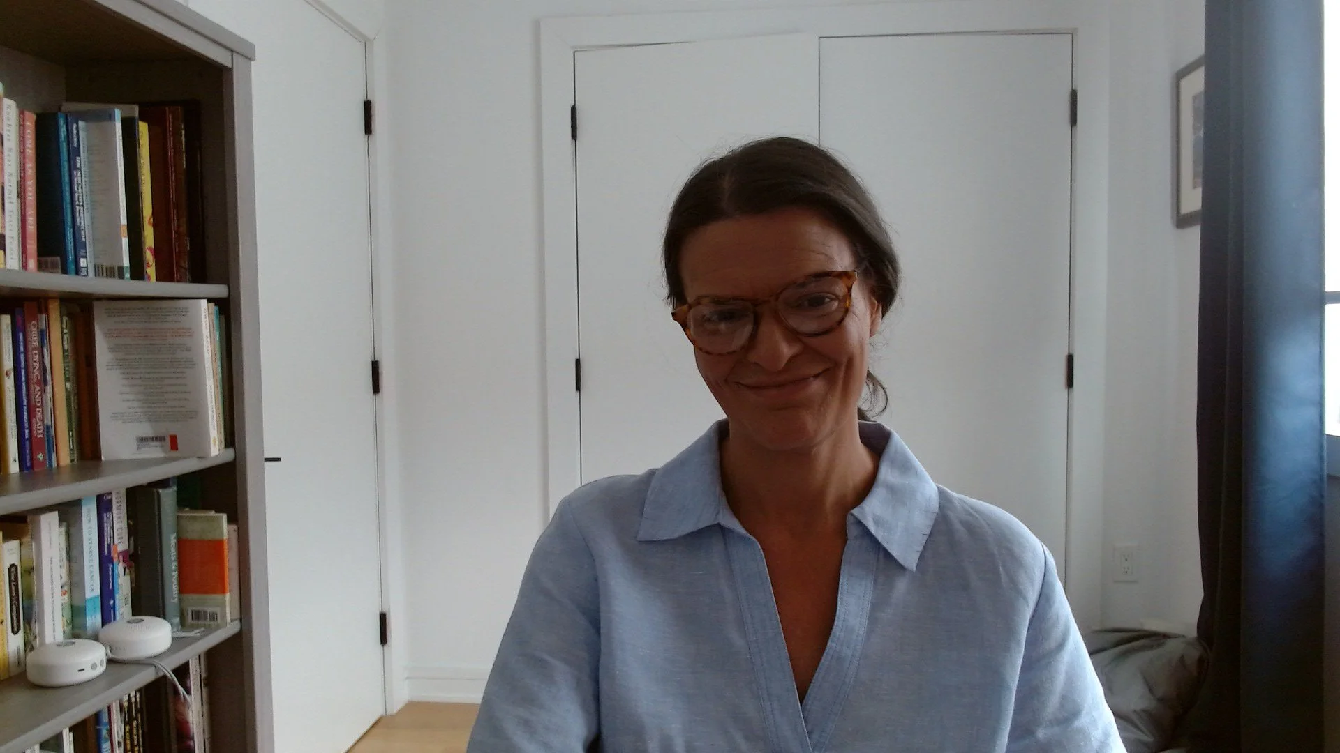 A woman with glasses and brown hair smiling at the camera. She is wearing a light blue shirt and is indoors, with a bookshelf filled with books on her left and a closed white door behind her.