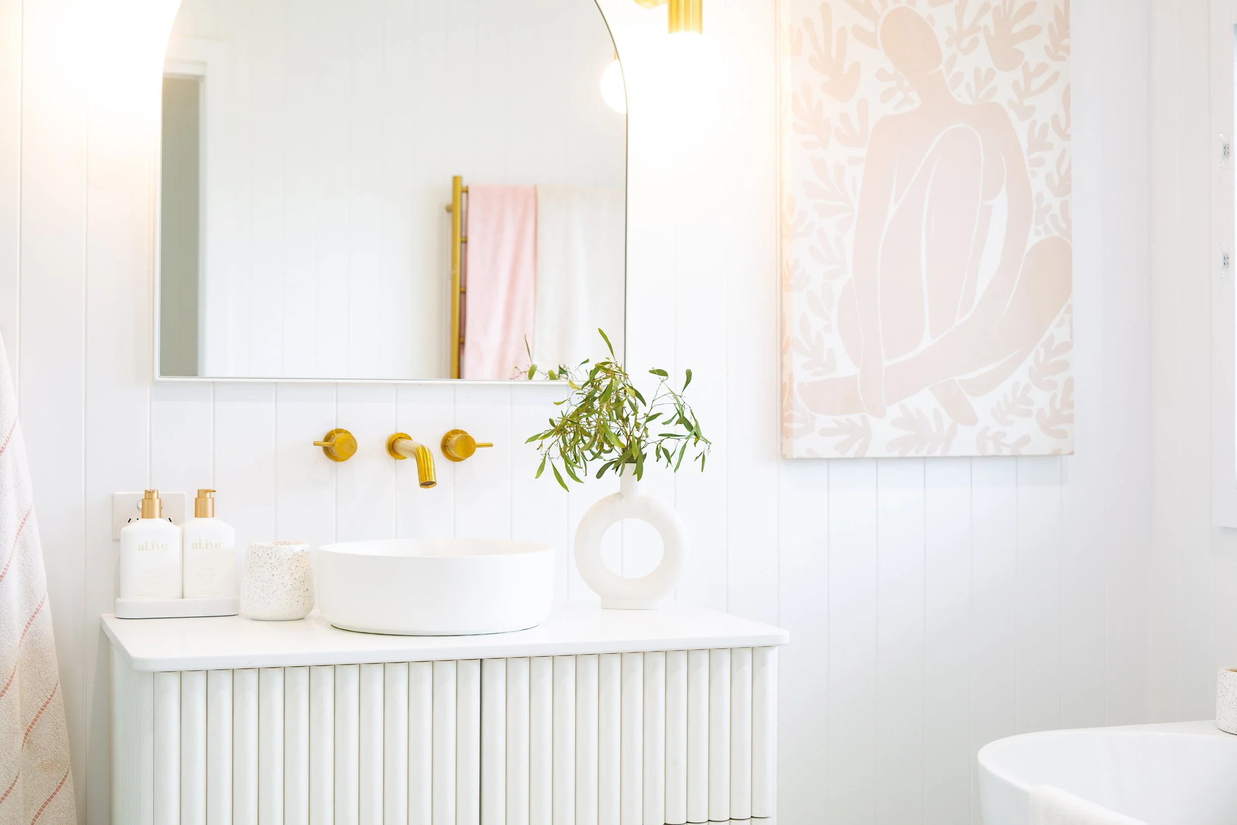 A minimalist bathroom with a white vanity, a white vessel sink, and a gold wall-mounted faucet. There are two white bottles labeled 'al:ive' on the vanity, along with a speckled container. A small green plant sits in a white vase. The mirror above the vanity reflects a towel rack with a pink towel and a window with bright light. To the right, a large canvas with a beige abstract design of a woman’s back hangs on the wall.