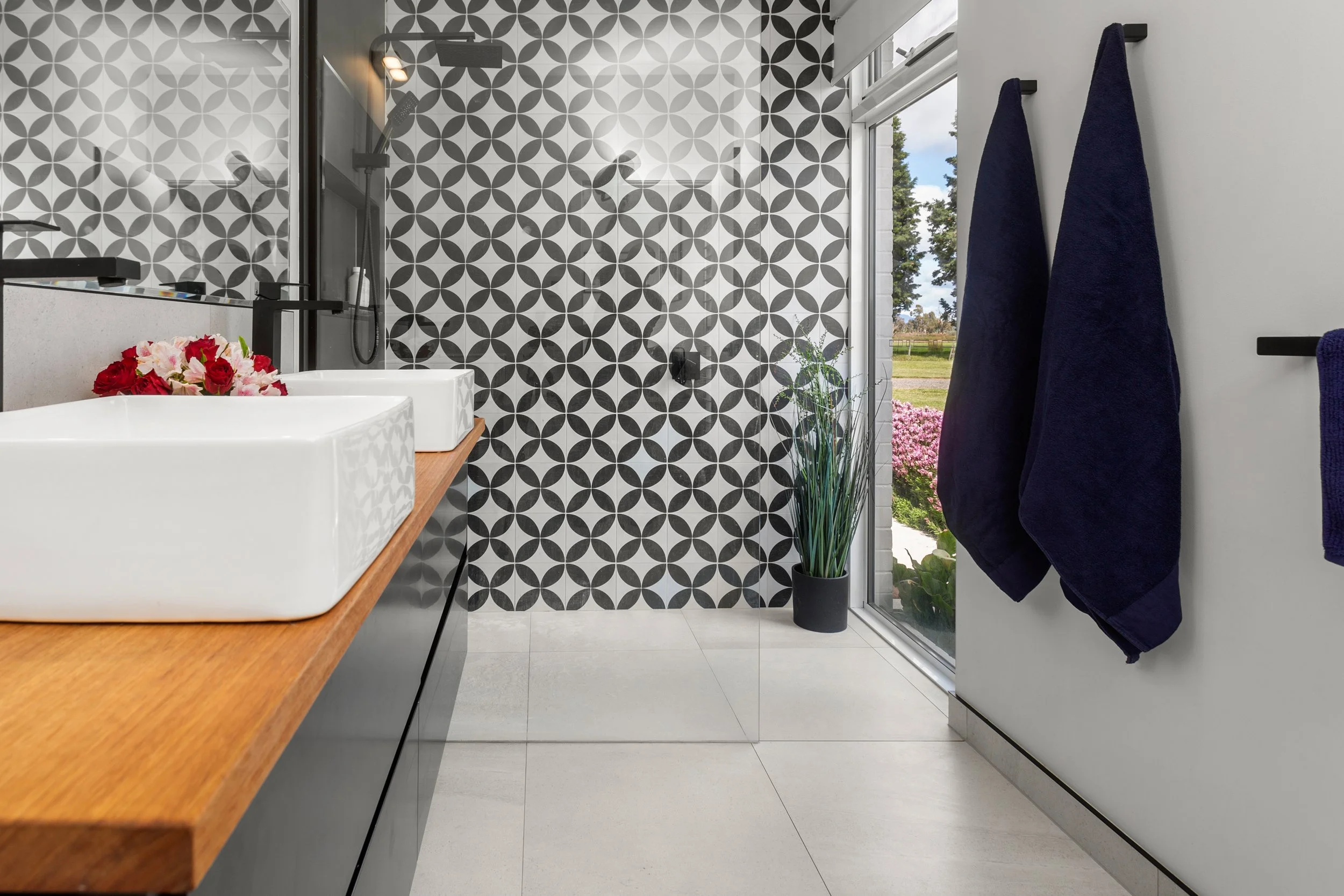 Modern bathroom with patterned black and white tile wall, white sink on a wooden countertop, dark towels hanging on the wall, and a large window showing outdoor greenery.