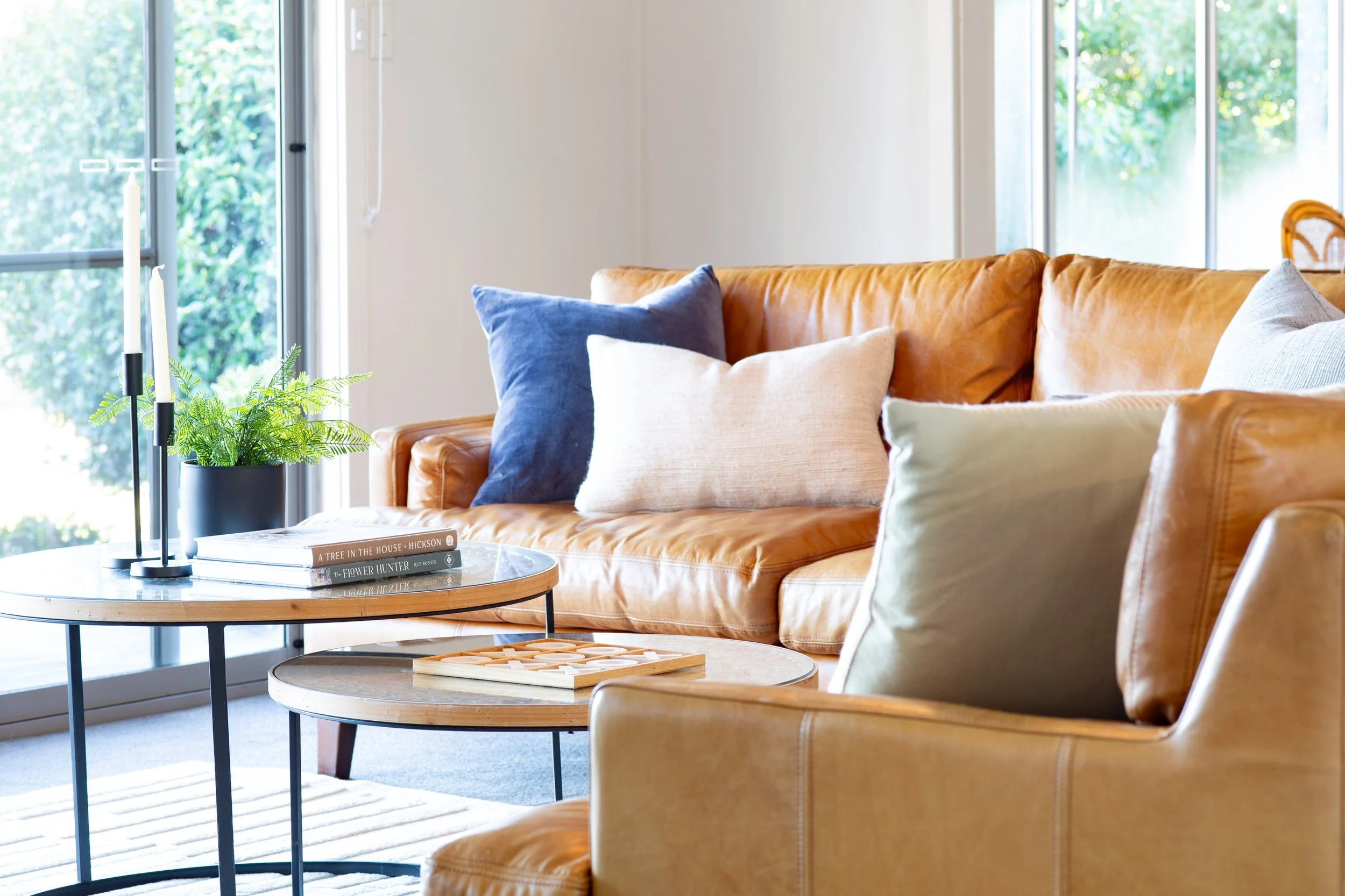 Living room with tan leather sofa, multiple pillows, a glass-top coffee table with books, a potted plant, and large windows showing greenery outside.
