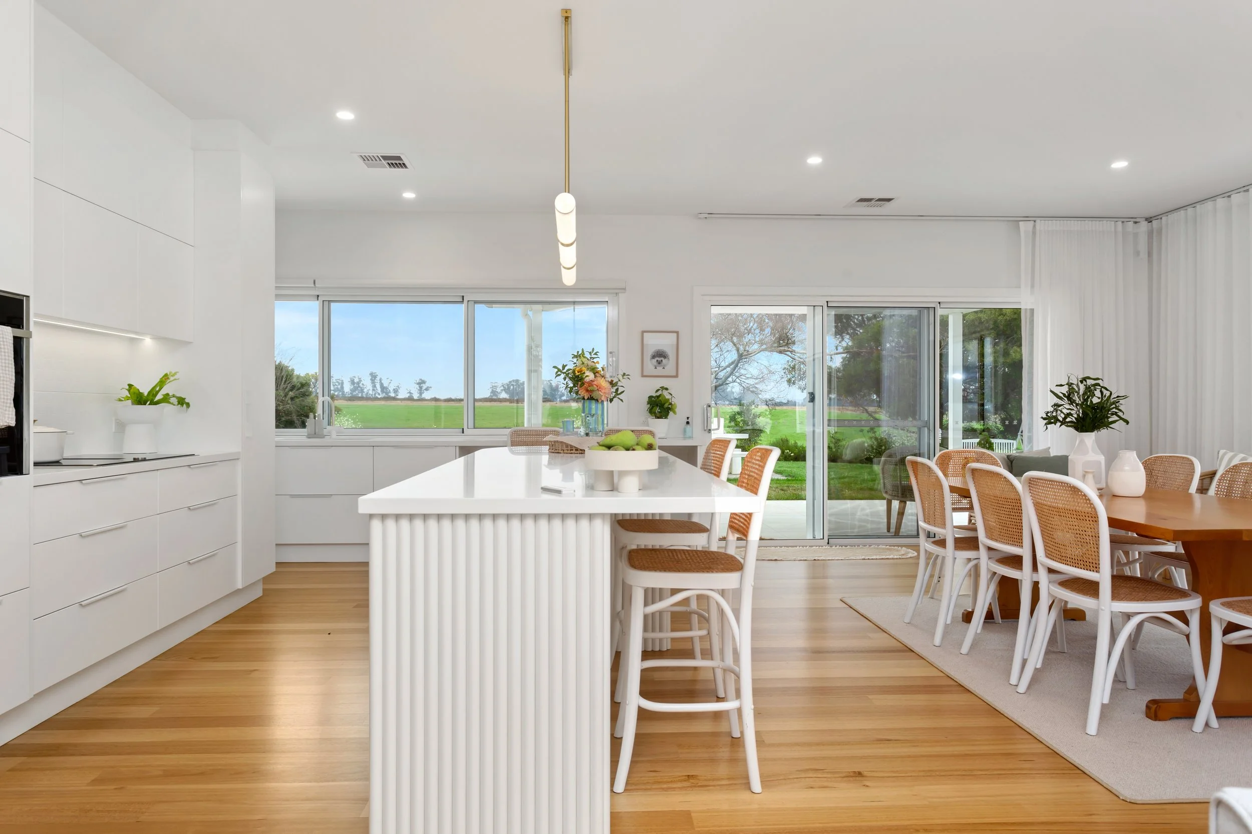 Bright and modern kitchen and dining area with large windows showing greenery outside, white cabinetry, a white island with vertical ribbed detailing, and a dining table with rattan-backed chairs, decorated with a floral centerpiece and vases.
