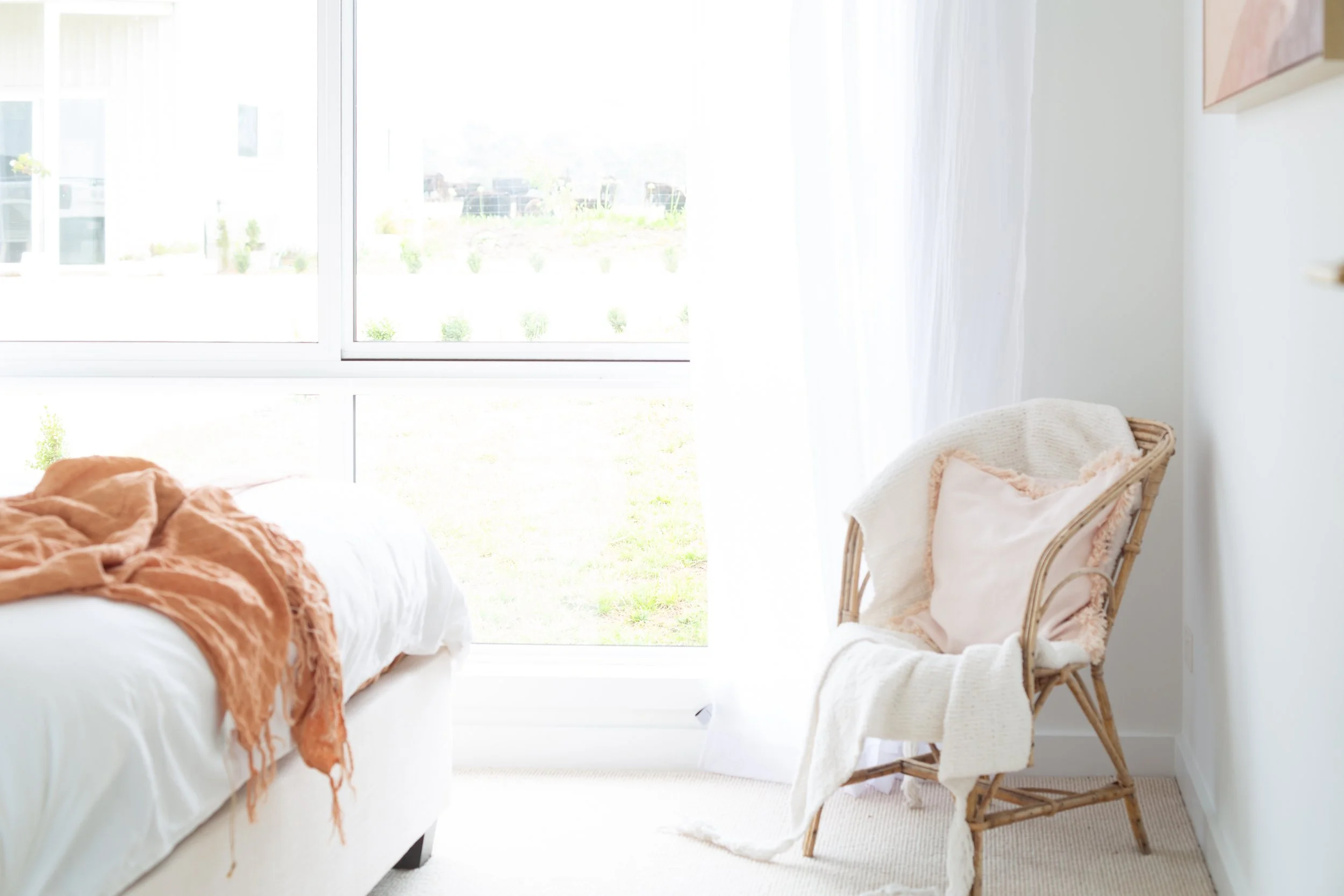 A cozy bedroom with a large window, a white bed with an orange blanket, and a wicker armchair with a blanket draped over it.