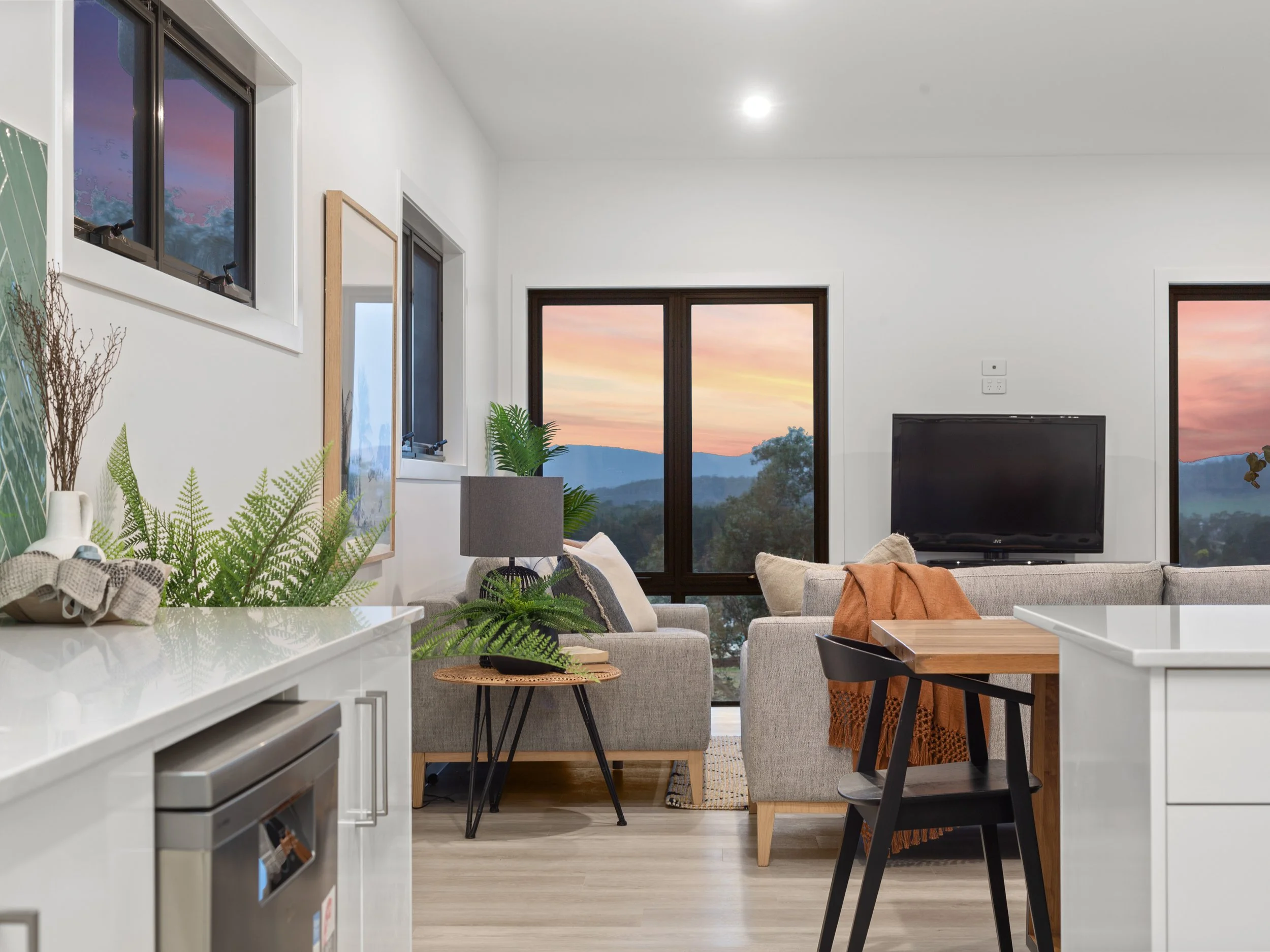 Modern living room with large windows showing a sunset view, neutral-colored furniture, green plants, and a flat-screen TV.