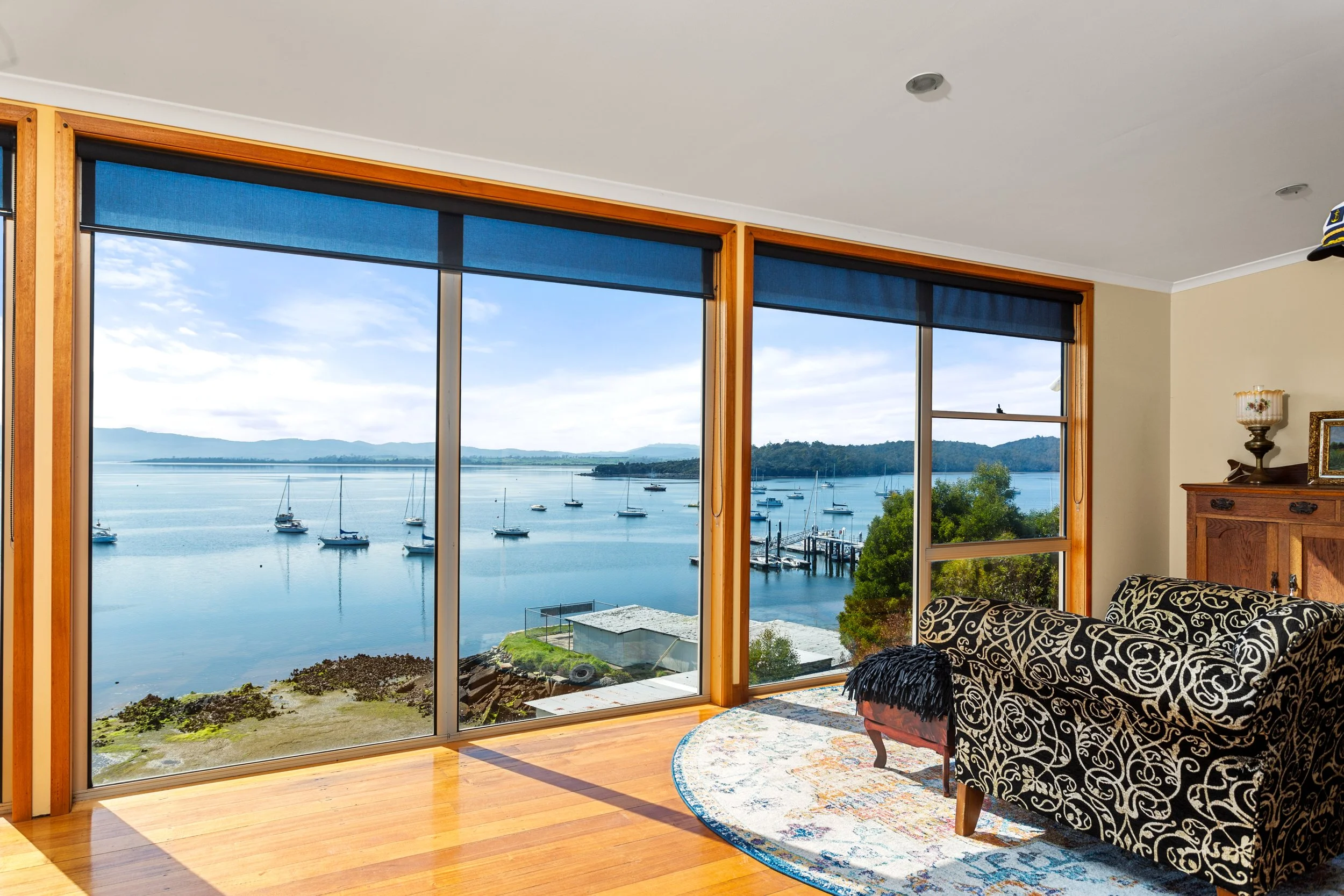 Living room with large front window overlooking a marina with boats and water, wooden flooring, patterned black and white armchair, and decorative lamp and picture frame on a wooden side table.