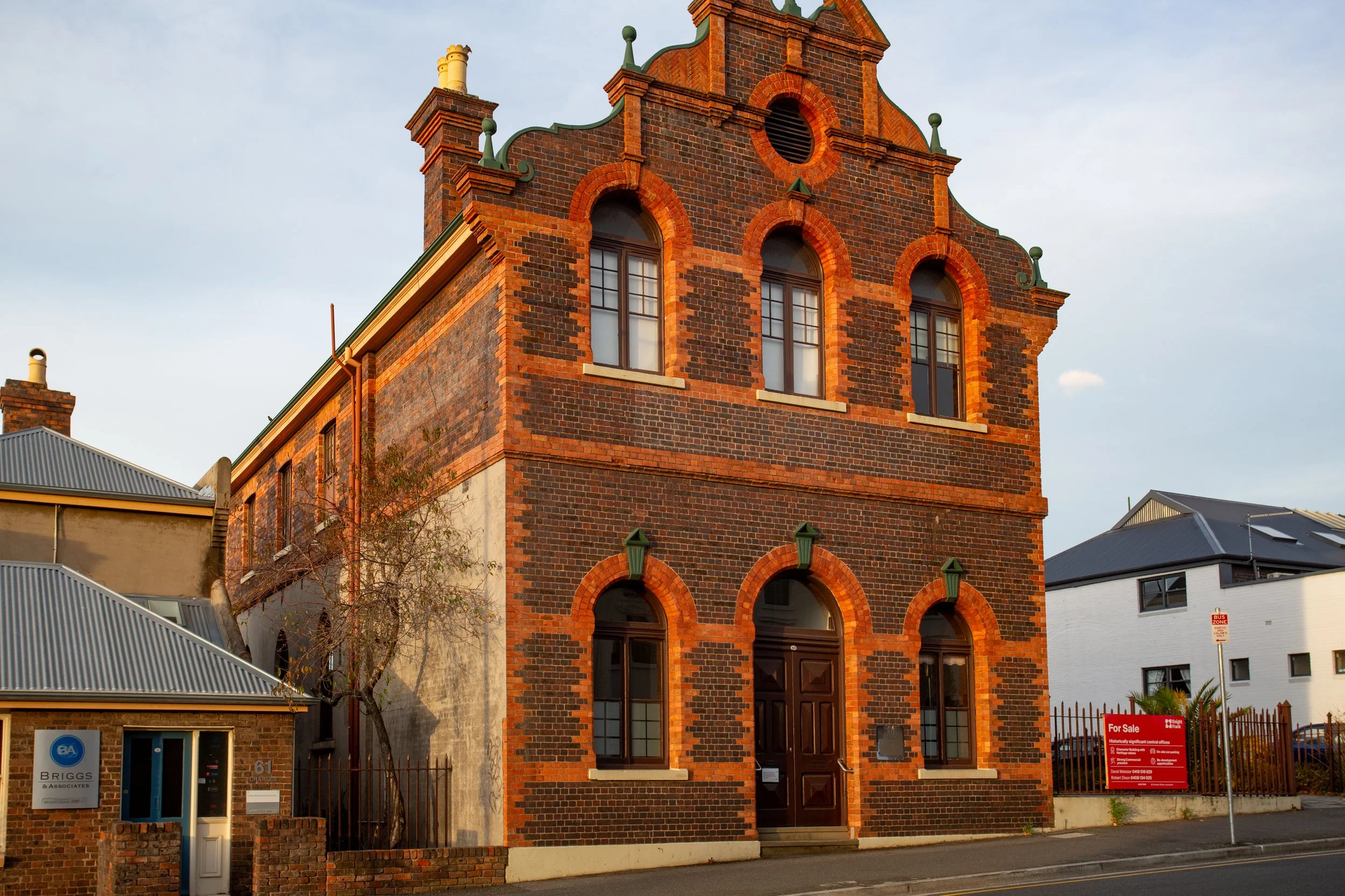 A historic brick building with arched windows and decorative elements on the upper part, surrounded by modern buildings, with a partly cloudy sky overhead.