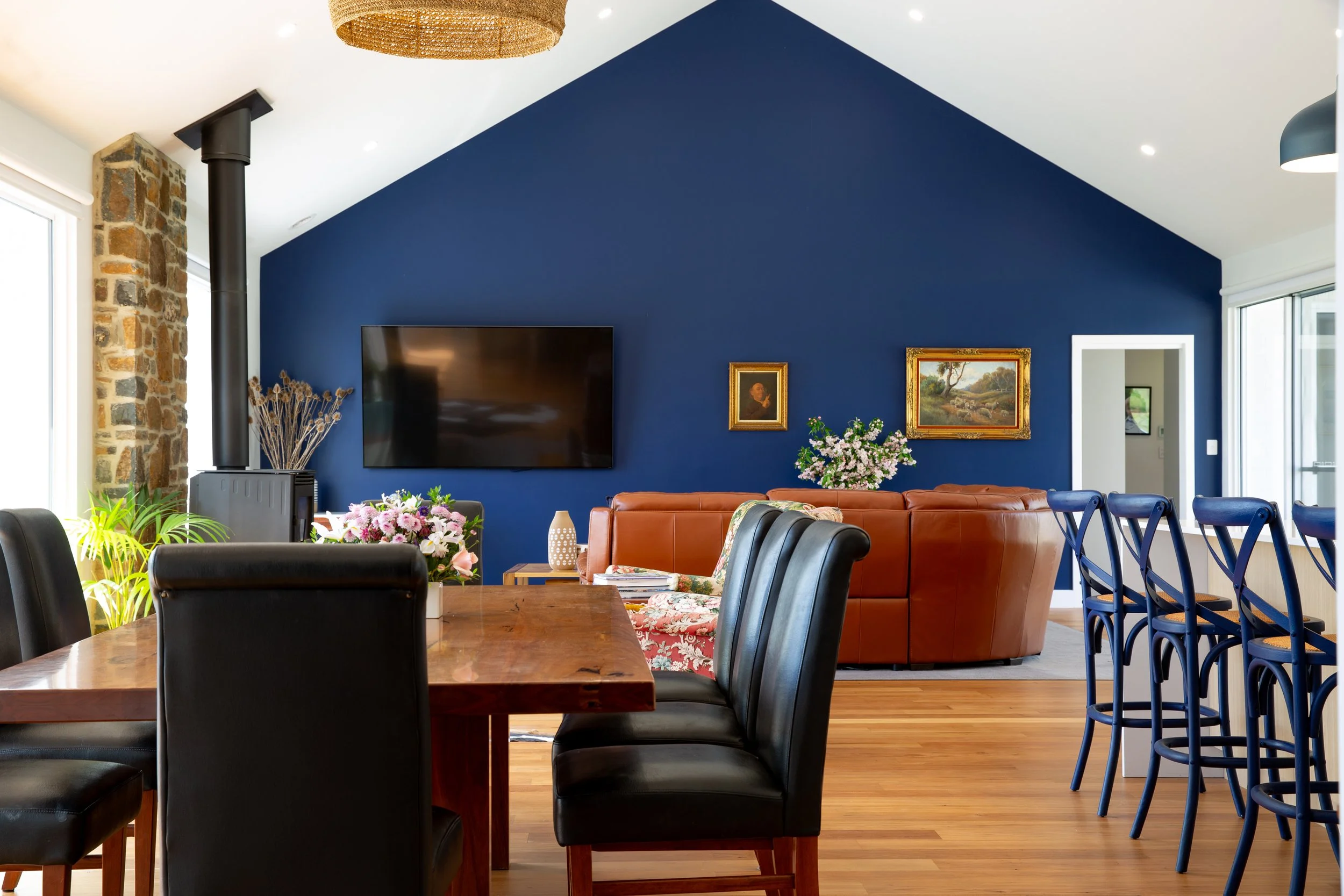 A spacious living and dining room with a blue accent wall, decorated with framed paintings and a flat-screen TV. There is a large wooden dining table with black chairs, a leather sofa, a bouquet of flowers, and large windows letting in natural light. The ceiling has a woven chandelier and a black stove pipe next to a stone wall.