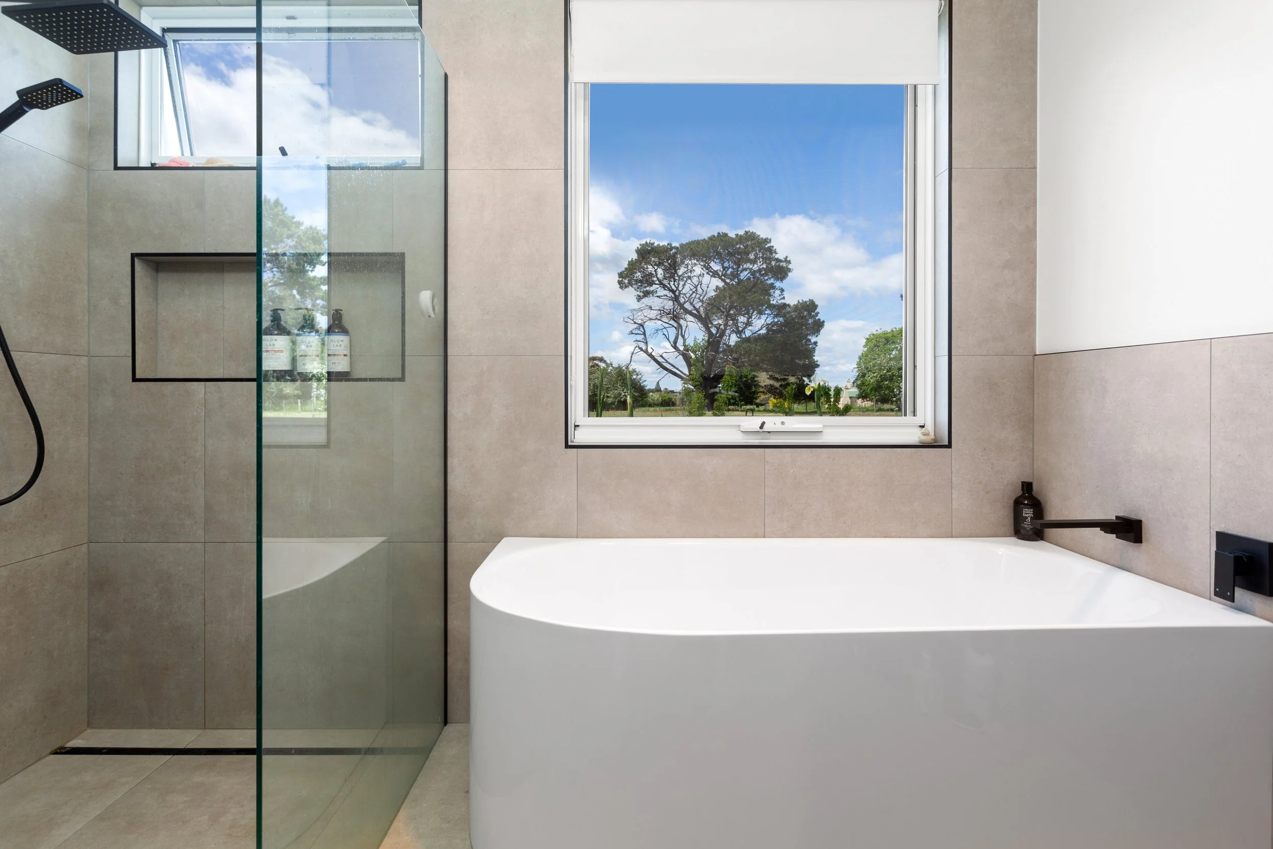 Modern bathroom with a freestanding bathtub, a glass shower with black fixtures, a large window showing trees and a blue sky, and neutral-toned tiled walls.