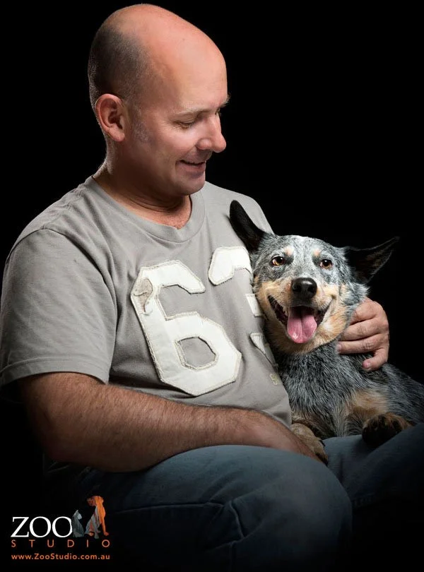 A man with a shaved head and wearing a gray T-shirt with a white '66' patch on it, smiling and looking down at his dog, a blue heeler with an open mouth and tongue out, sitting on his lap against a black background.