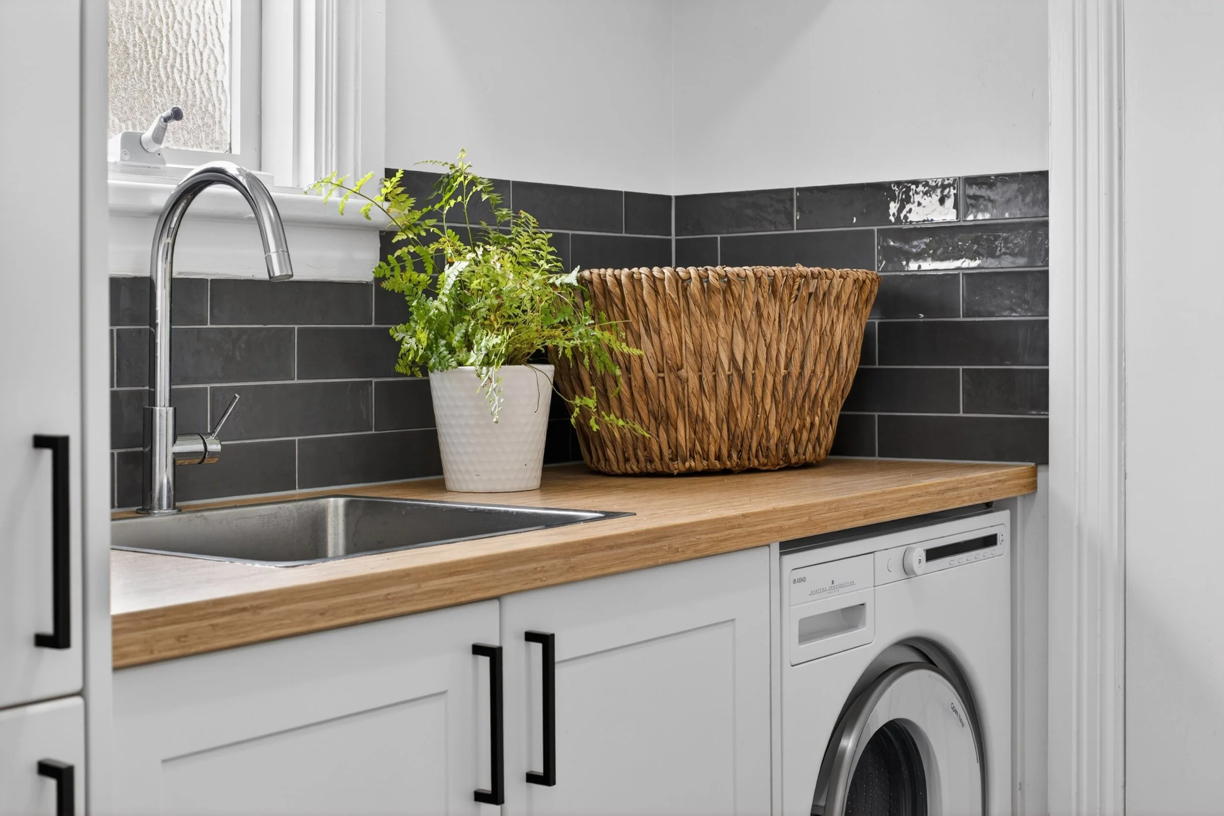 A laundry area with a kitchen countertop featuring a kitchen faucet, a potted green fern plant, a wicker basket, and a washing machine in a white cabinet base.