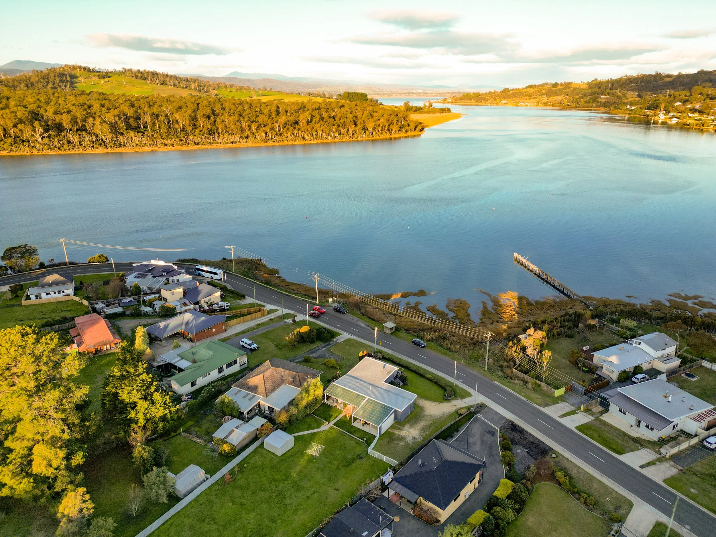 Aerial view of a suburban neighborhood by a large river, with houses, streets, trees, and a dock extending into the water, surrounded by green hills and mountains in the background.