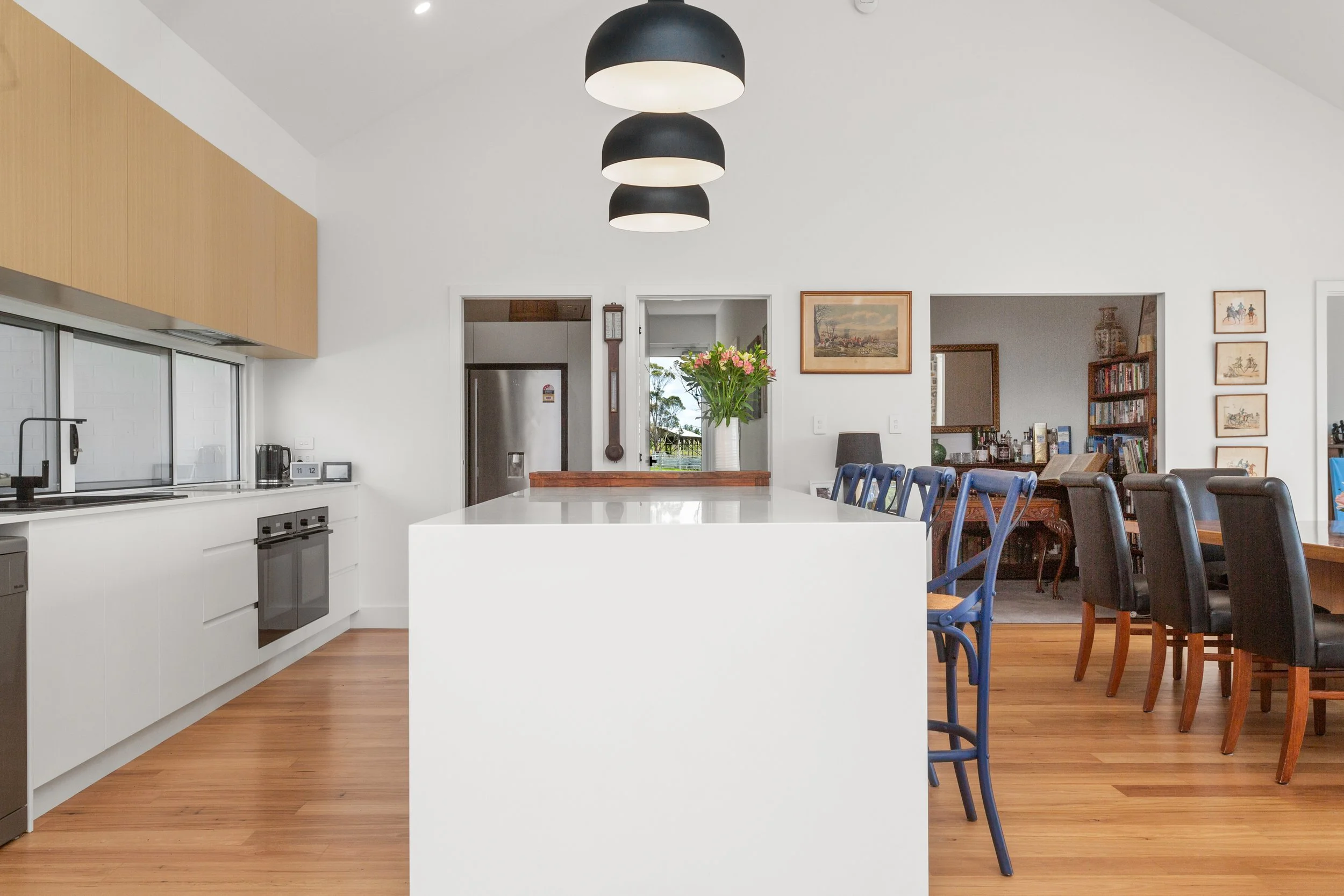 Modern kitchen and dining area with white island, black and blue chairs, wooden floors, wall art, and a vase of pink flowers on the island.