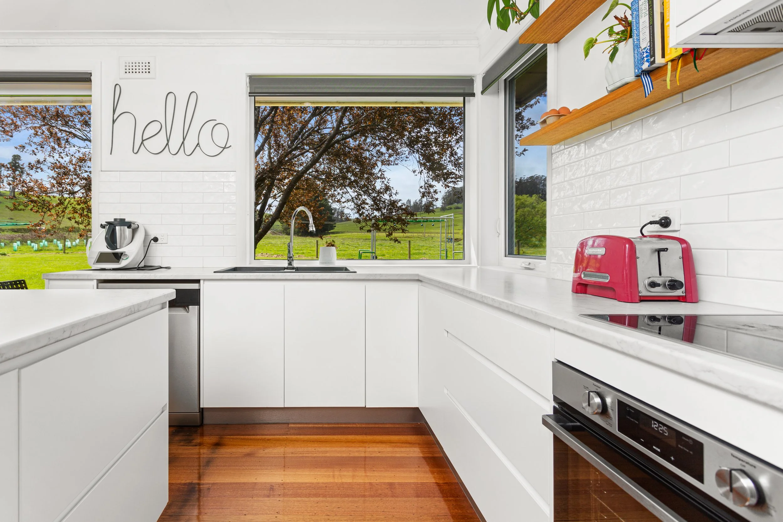 Modern kitchen with white cabinets, marble countertops, and large window overlooking green fields and trees.