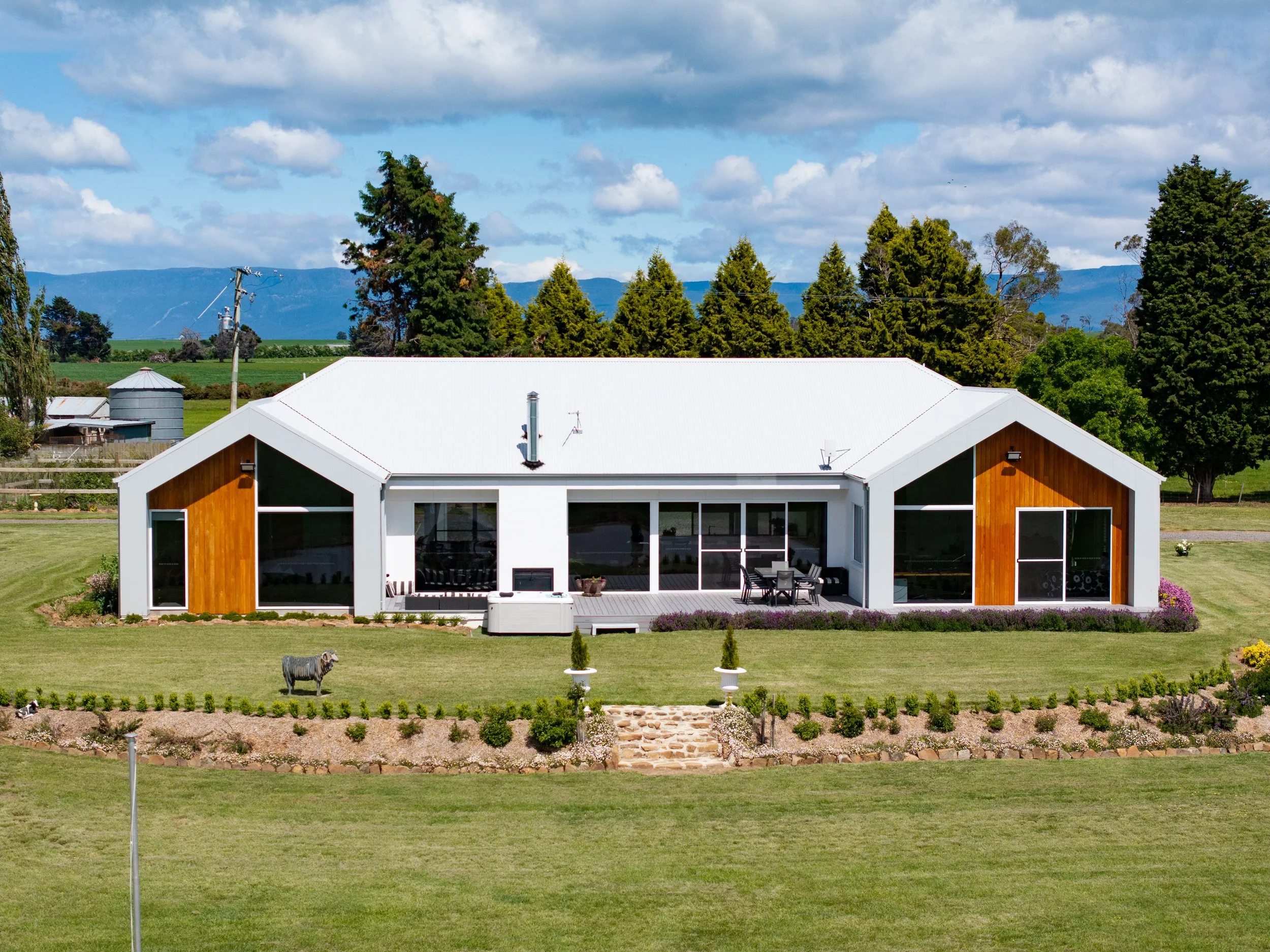 A modern white house with wooden accents and large glass windows, surrounded by a green lawn, bushes, and garden beds, with a backdrop of trees and mountains under a partly cloudy sky.