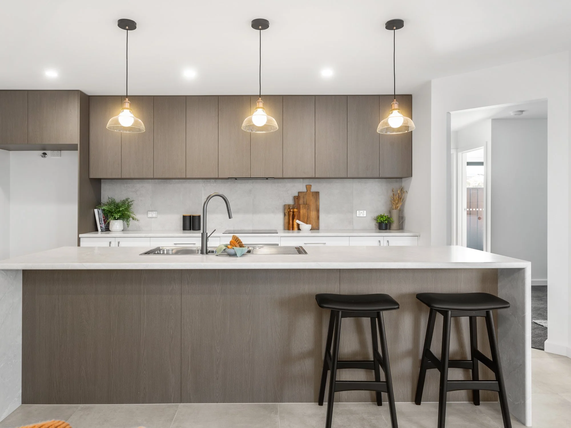 Modern kitchen with beige cabinets, white countertops, a waterfall island, black bar stools, pendant lights, plants, and kitchen utensils.