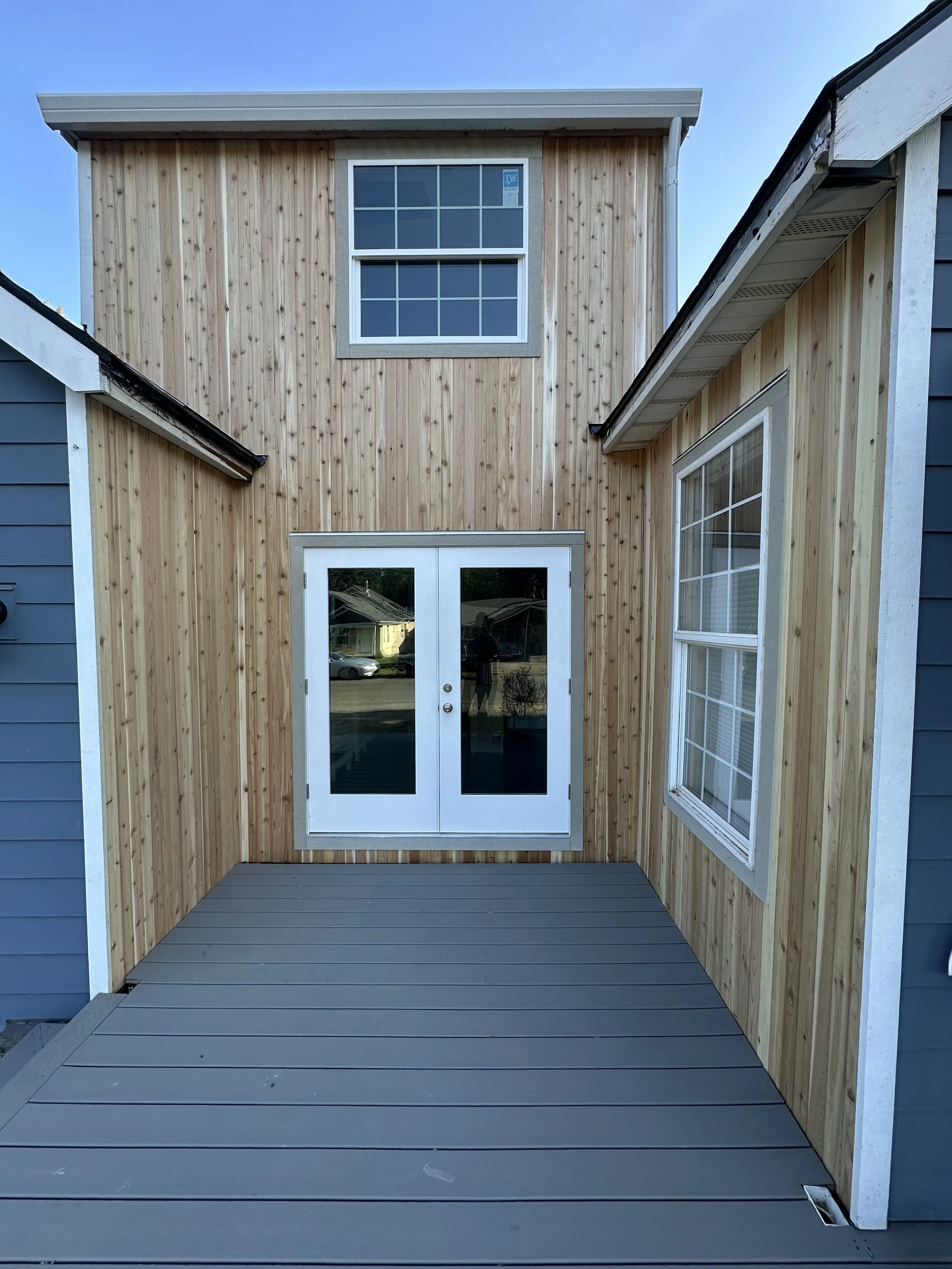 Small outdoor patio with grey decking, wooden siding on walls, and white-framed glass double doors, surrounded by neighboring houses under a clear blue sky.