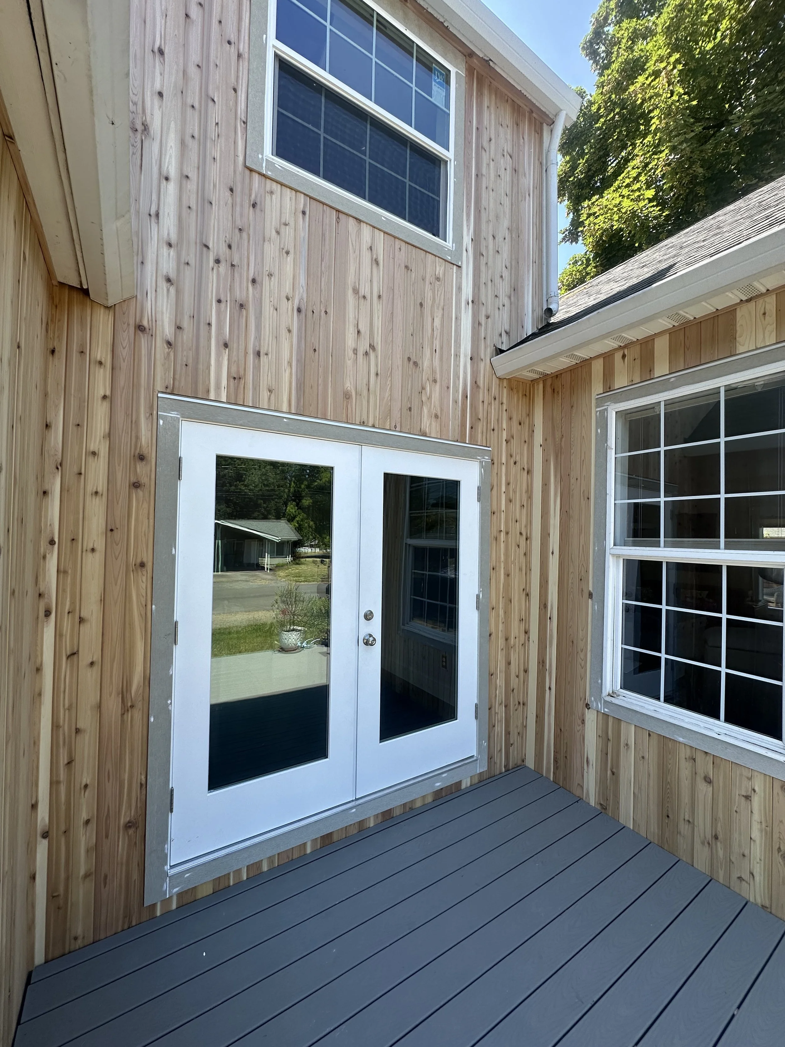 A backyard patio with gray wooden flooring, a wooden exterior wall, and a white door with glass panels. There are windows with white frames and a view of a green lawn and houses in the background, with a large leafy tree on the right.