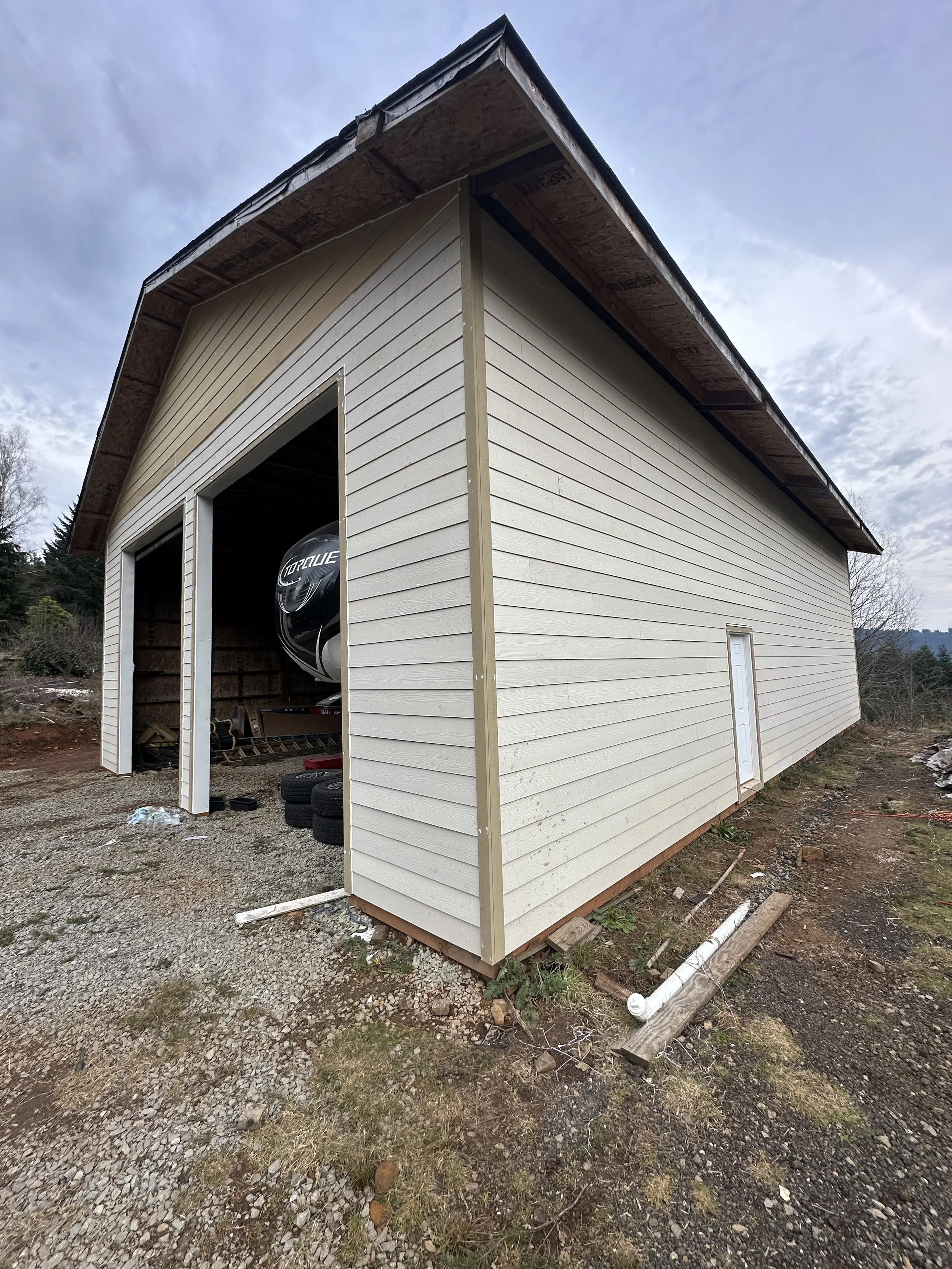 Photo of a building under construction with beige siding, an open garage, and construction materials on the ground.