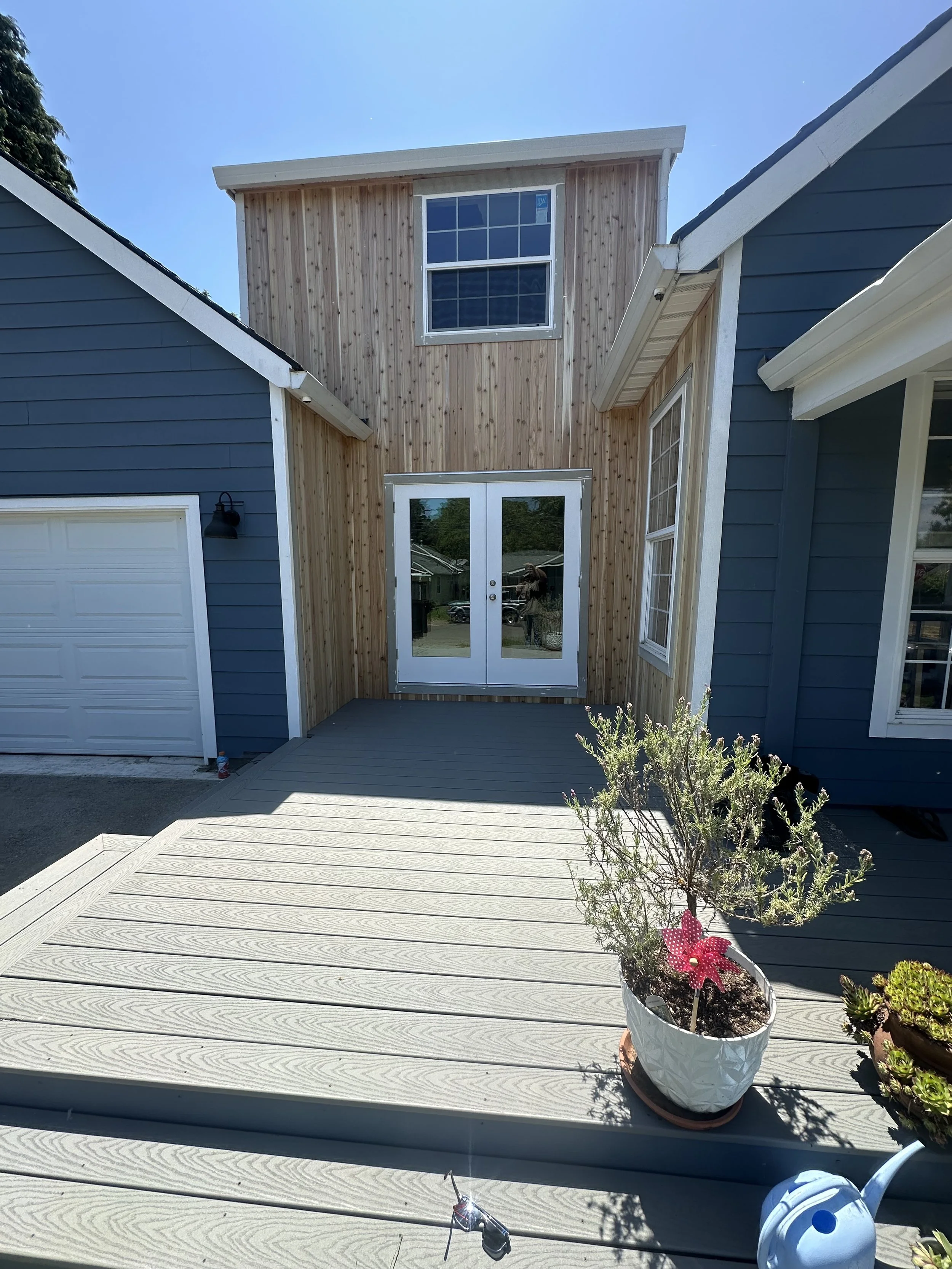 View of a house with a partially constructed upper story, featuring blue and natural wood siding, a glass door on the porch, and potted plants on the deck under a clear blue sky.