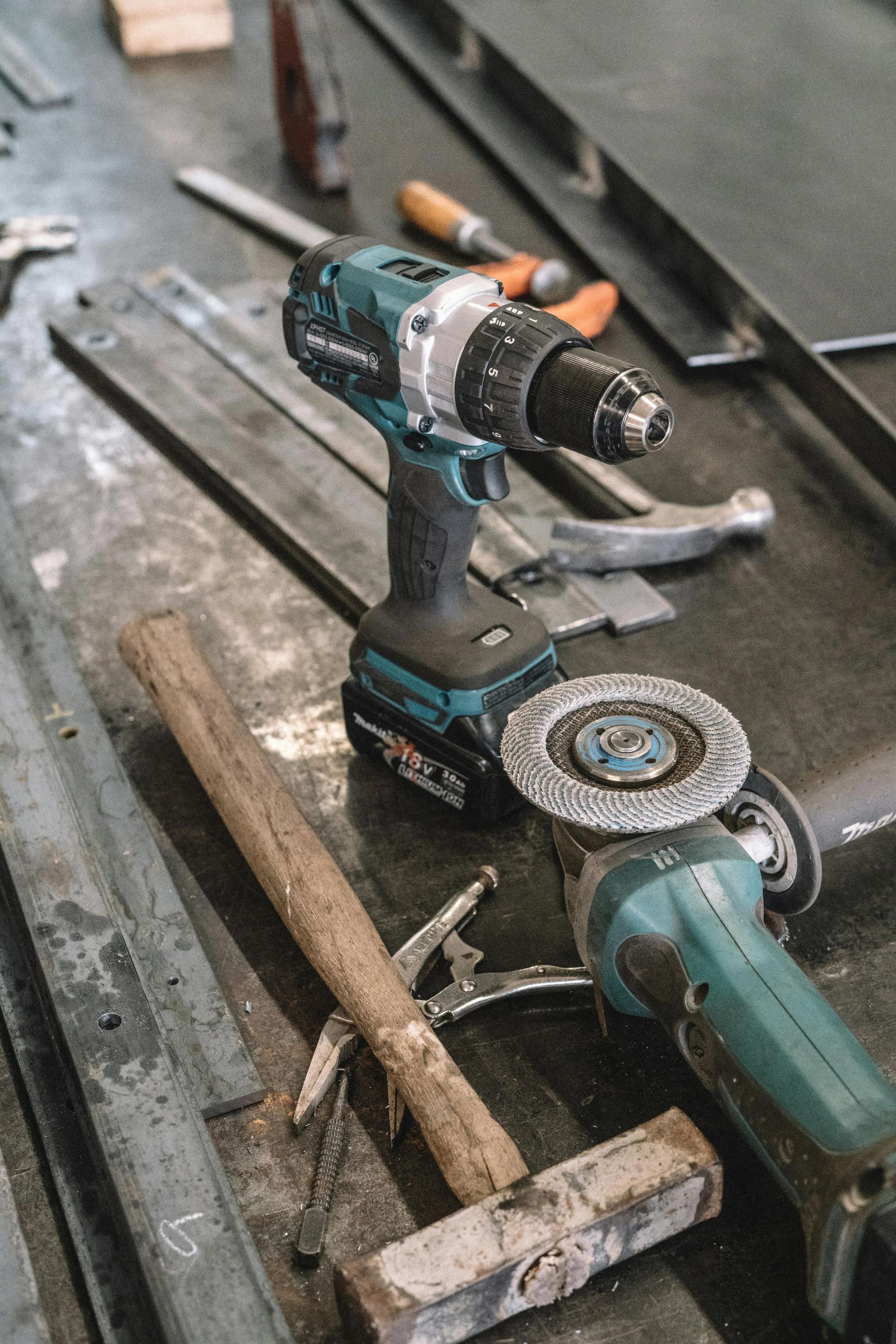 A cordless power drill resting on a workbench, with a wire brush attachment and a grinding disc nearby, in a workshop setting with tools and clamps.