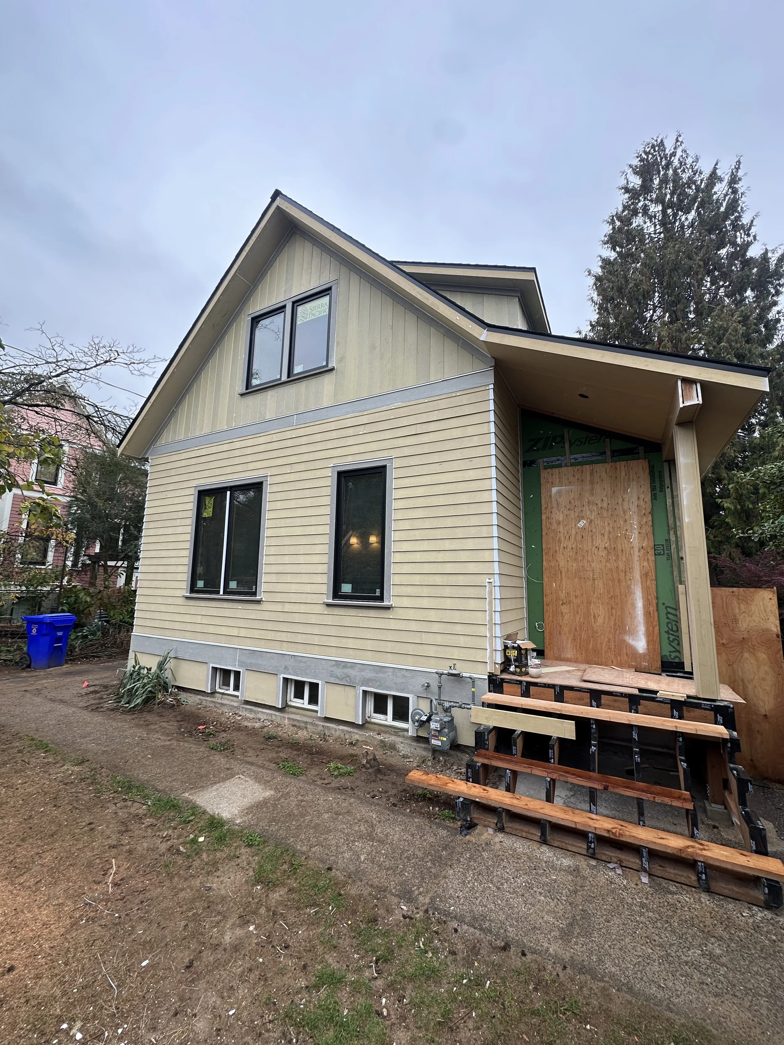 A house under construction with yellow siding, multiple windows, and a partly finished entrance with stairs and construction materials nearby. The yard has dirt and some small plants.