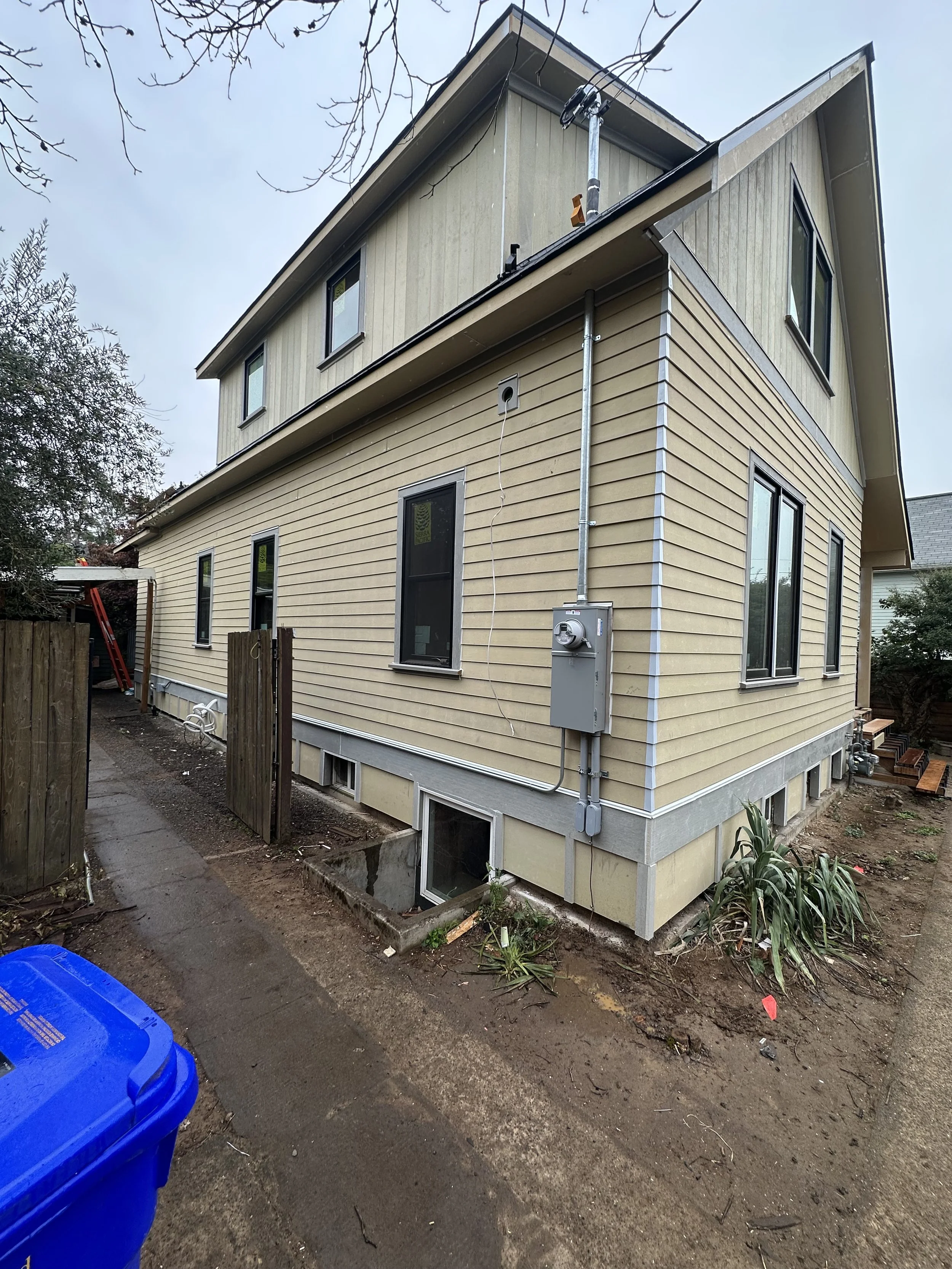 A multi-story house under construction or renovation with beige and greenish siding. There are several windows, a gray electrical box on the exterior, and a drainage pipe. The yard is bordered by a wooden fence with some plants and construction mater